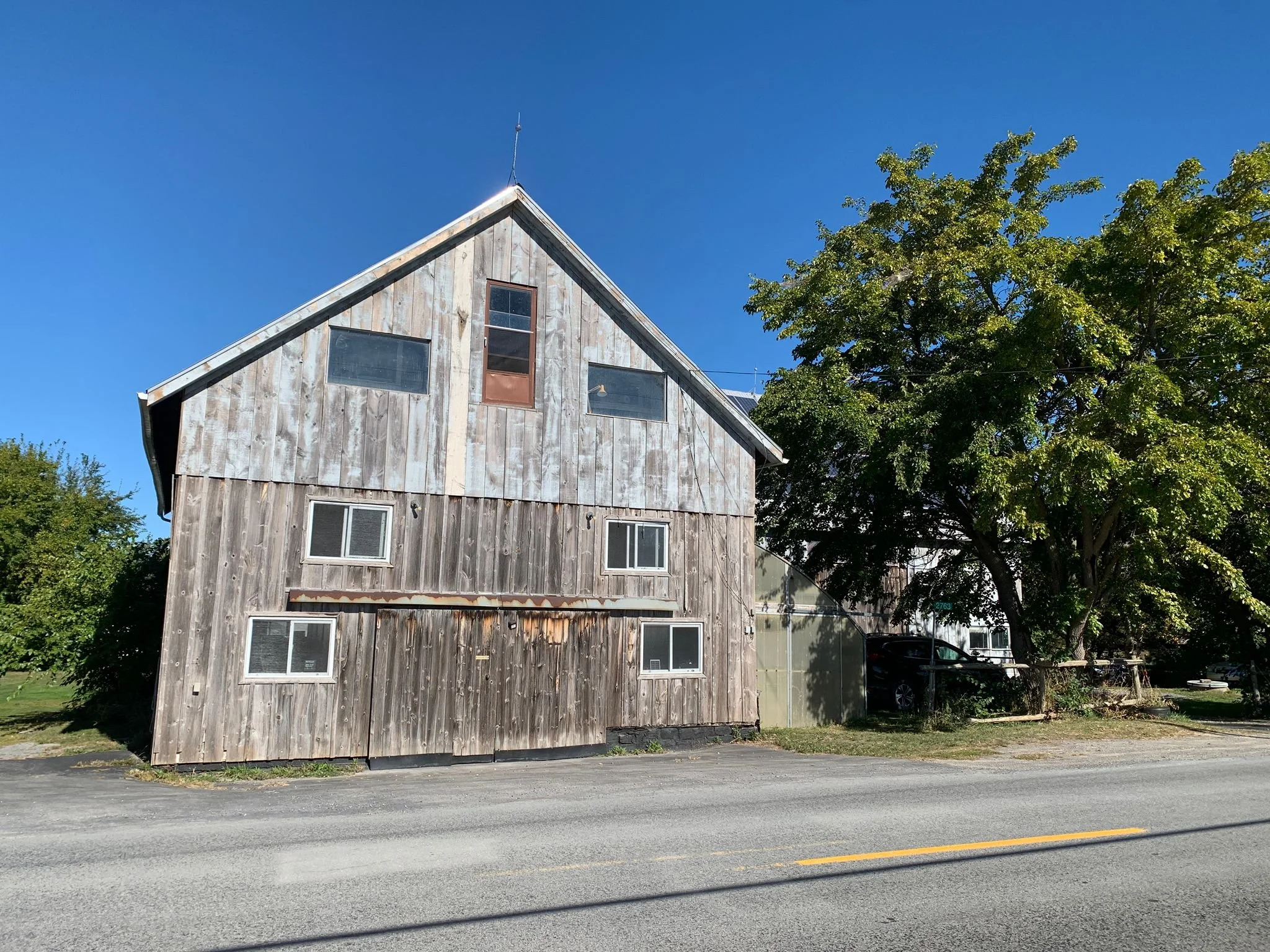 A two-story building with a weathered wooden exterior, several small windows, and a pitched roof, situated next to a large leafy tree under a bright blue sky.