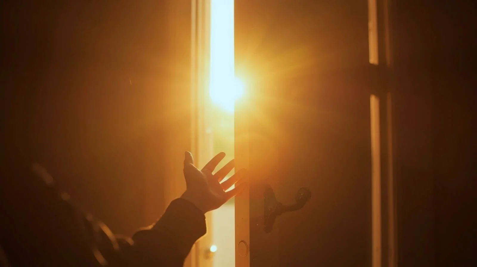 A hand reaching out to open a door as sunlight shines through the gap, creating a warm glow.