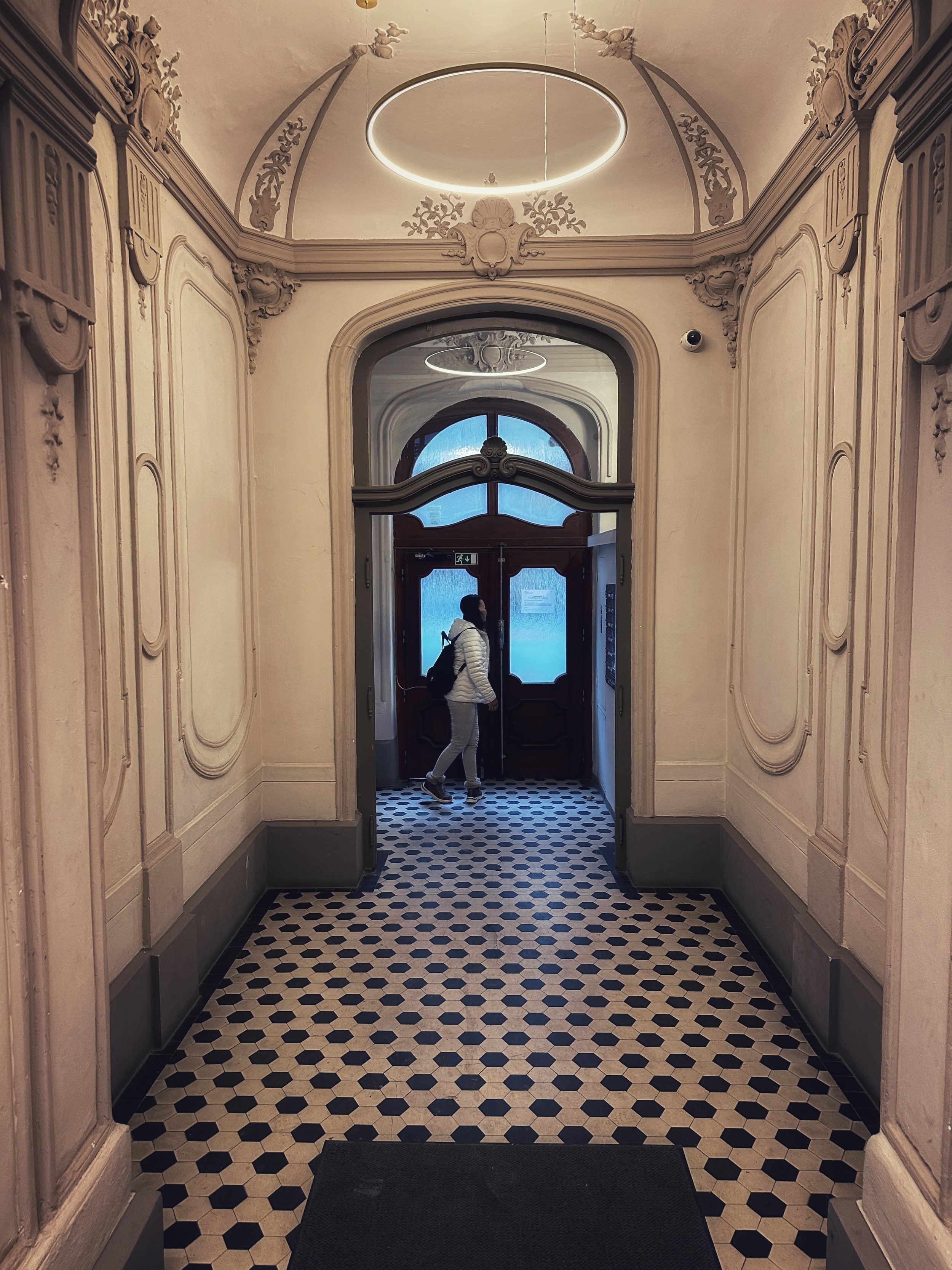 Interior corridor of a historic building in Prague, with patterned tile floor and a person walking toward a glass door.