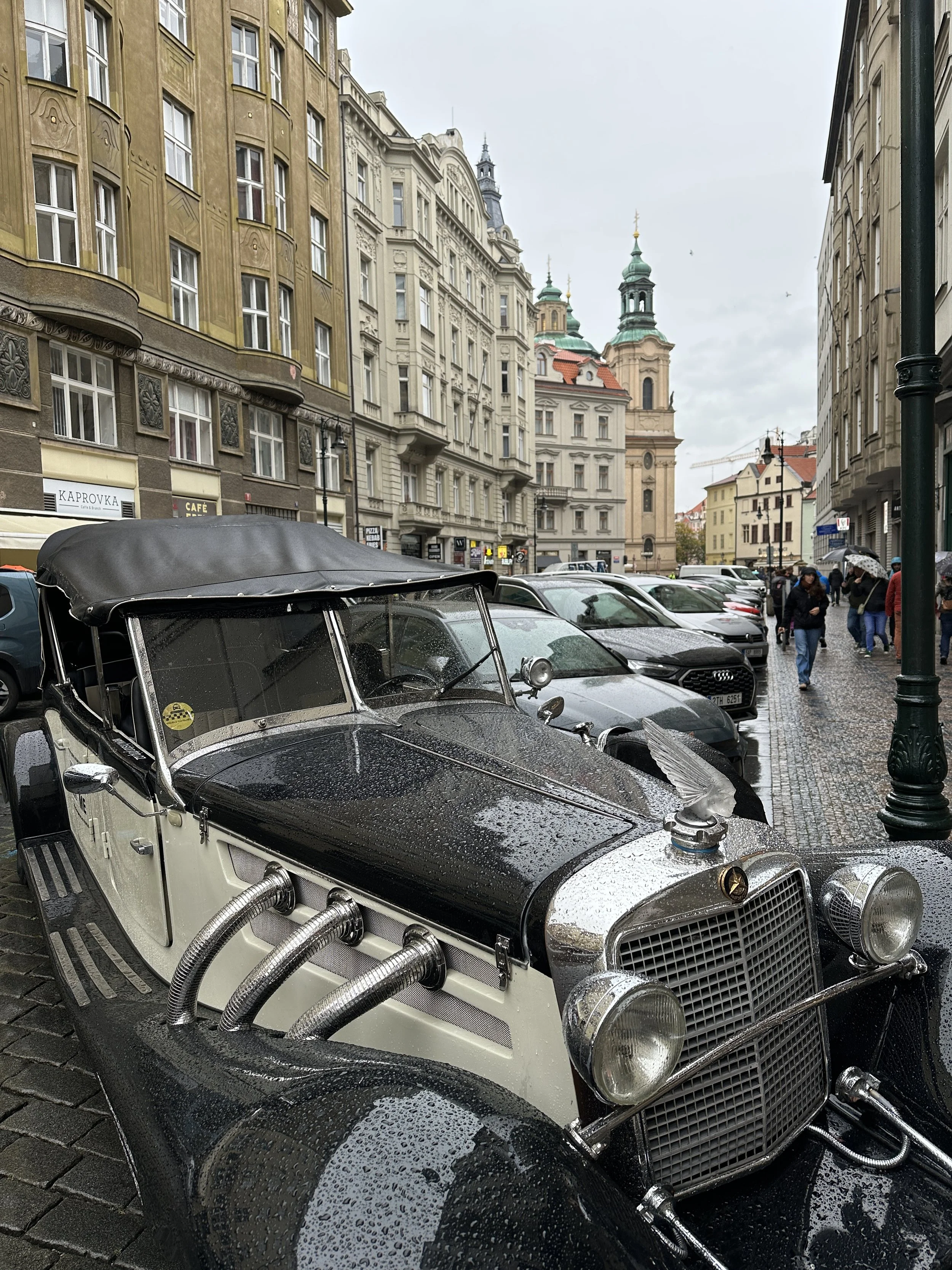 Vintage car parked on a wet street in Prague, with historic buildings lining the background.