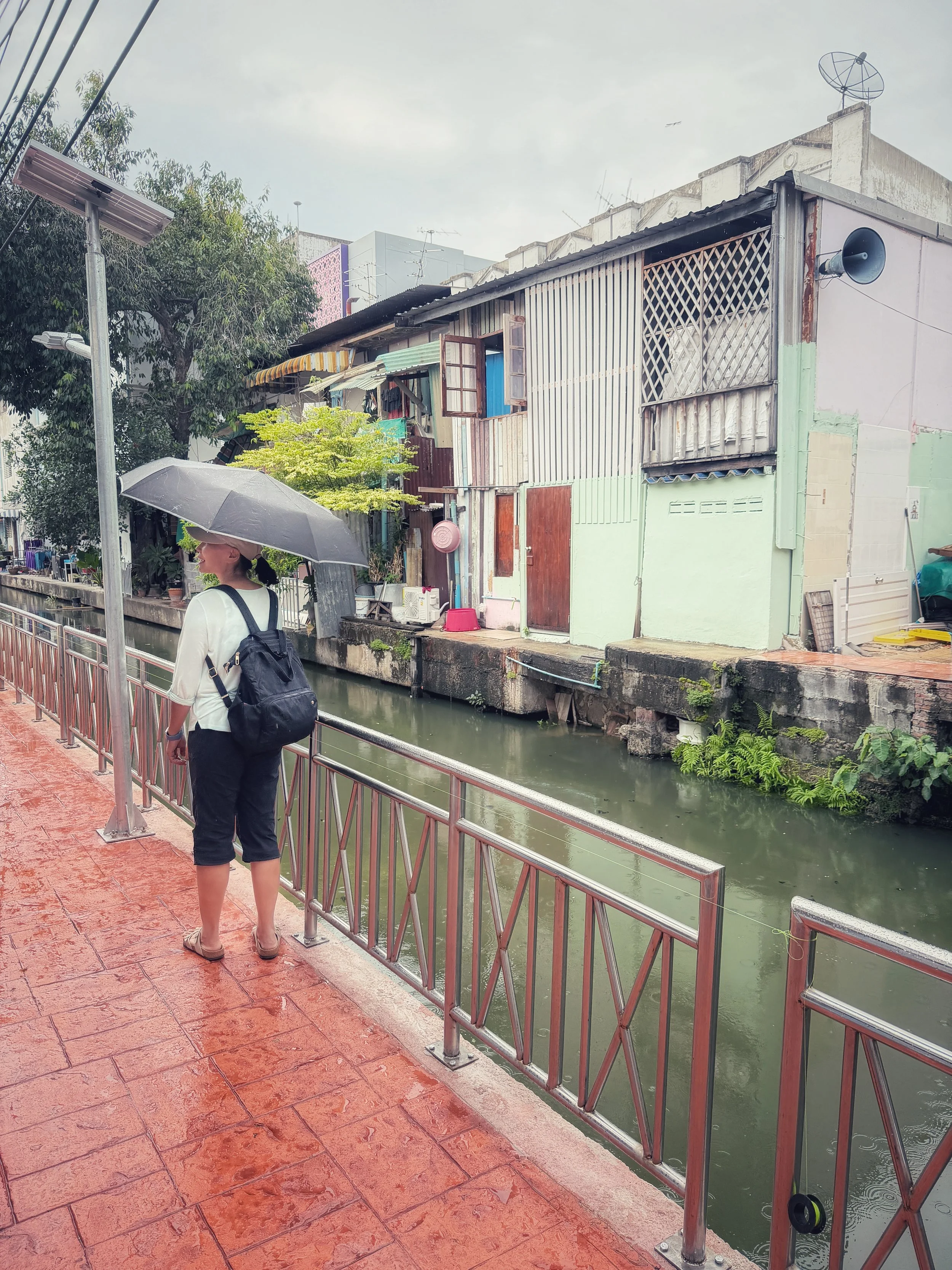 Person with an umbrella walking beside a Bangkok canal on a wet, overcast day.