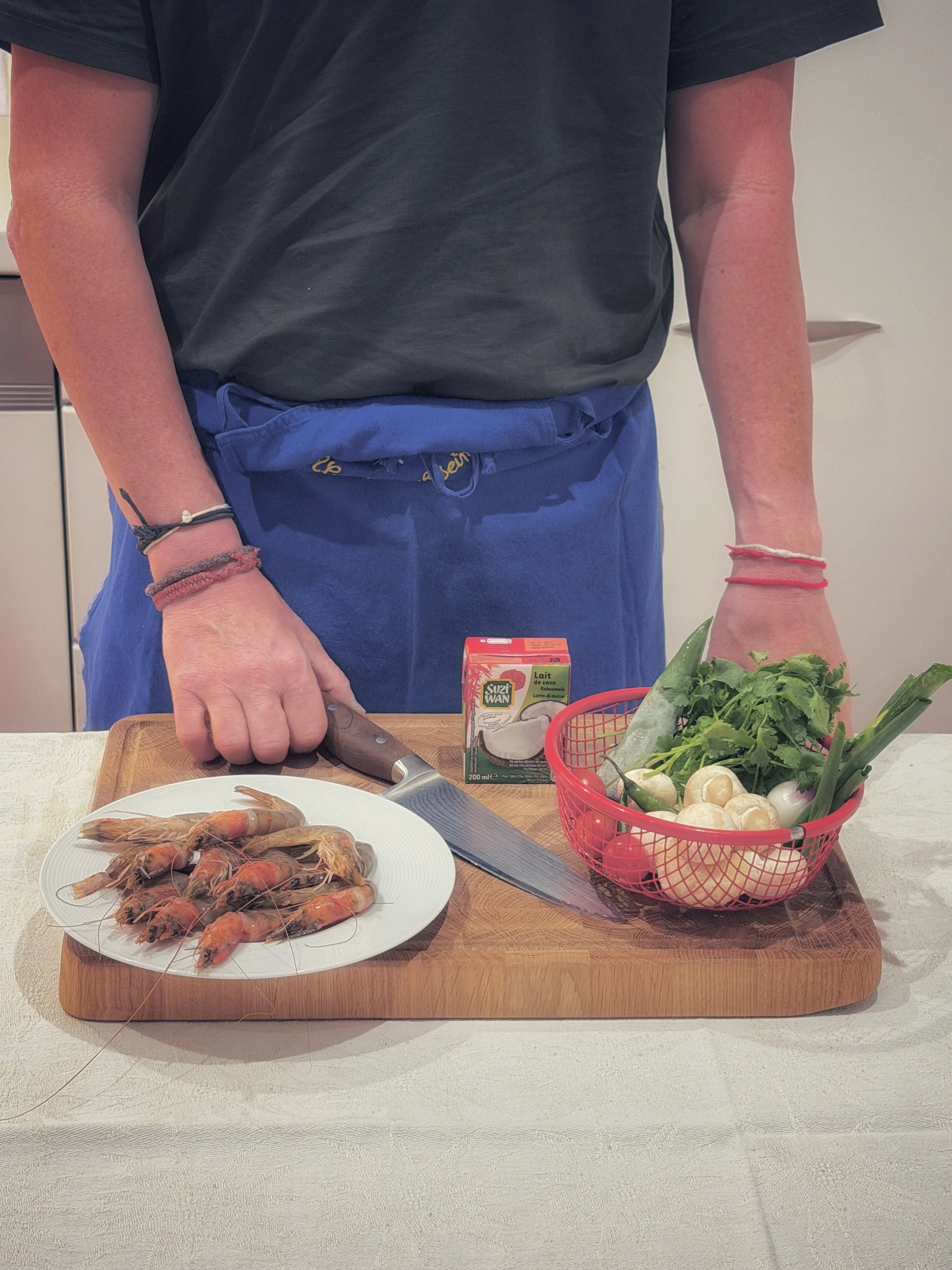 Preparing a Thai dish at home before travelling to Thailand, with shrimp, fresh herbs, vegetables, coconut milk, and a chef's knife arranged on a wooden board.