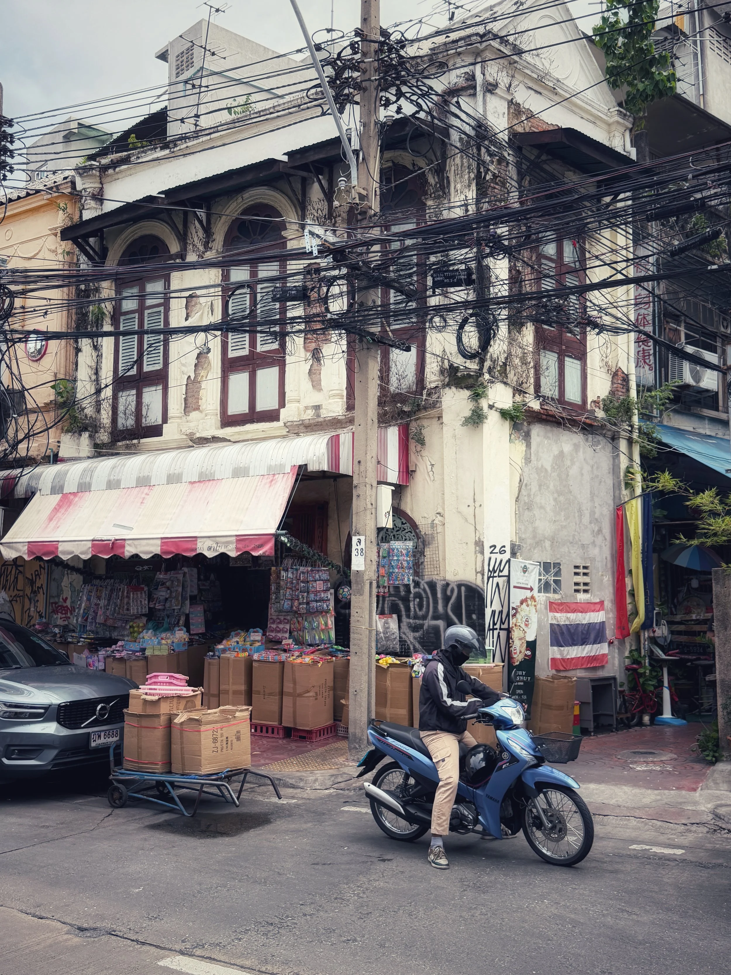 Weathered Bangkok building crossed by dense electrical wires, with a motorcyclist passing in front.