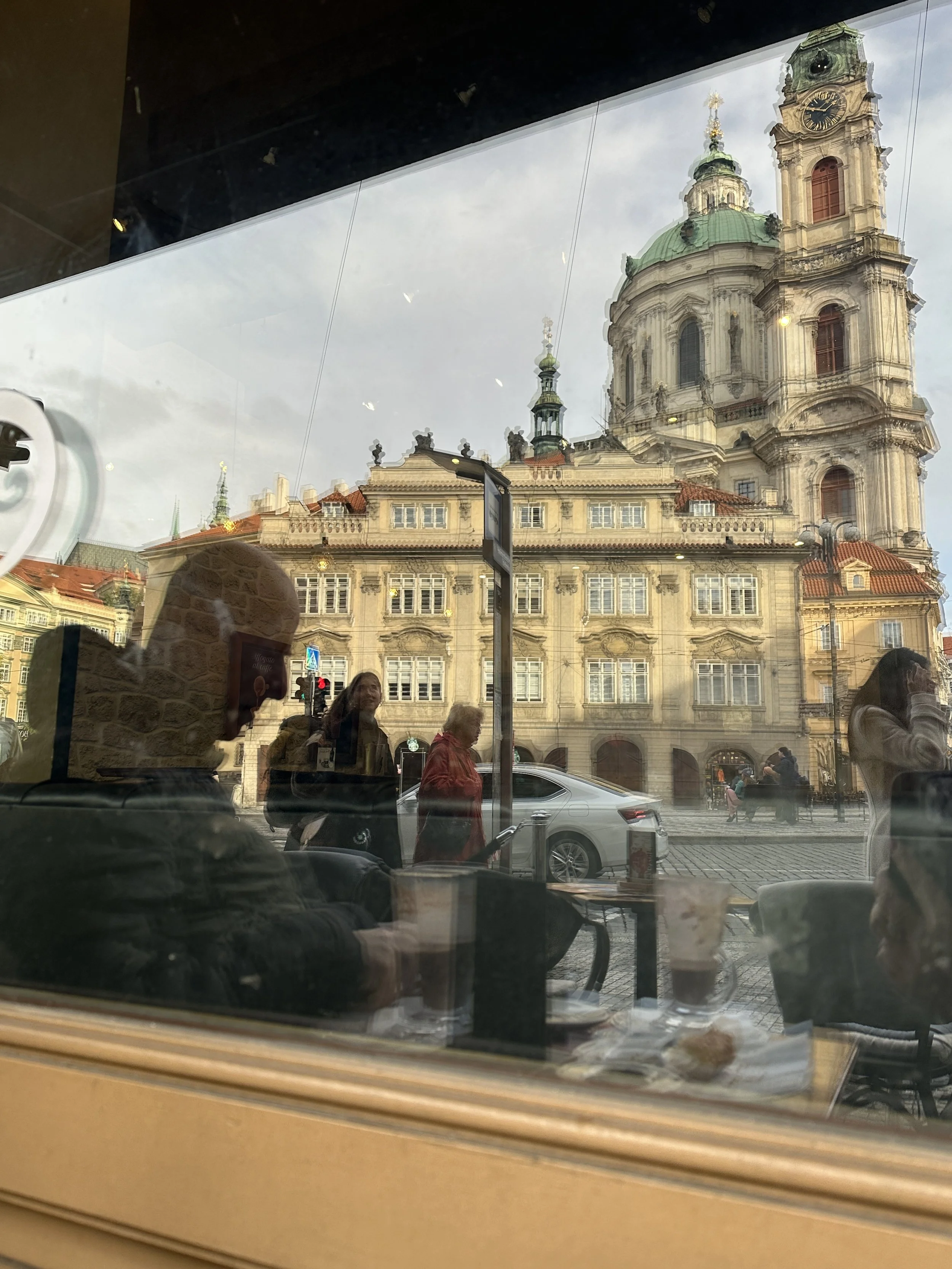 Reflection of a Prague church seen through a café window, people seated inside and city life outside