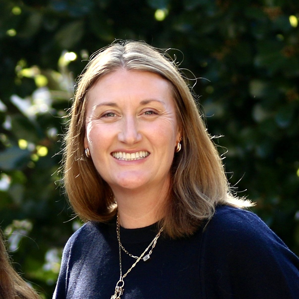 A smiling woman with shoulder-length light-brown hair, wearing earrings, layered necklaces, and a blue top, stands outdoors against a backdrop of greenery.