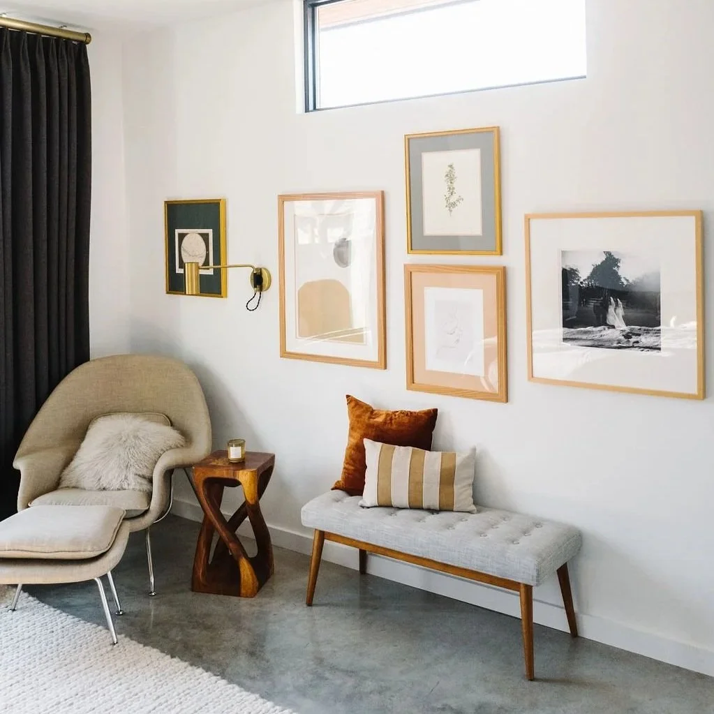 A stylish living room corner with a gray upholstered bench, pillows, and a modern gallery wall above. The art is framed in natural wood and thin gold frames, mixing black and white and minimalist prints.
