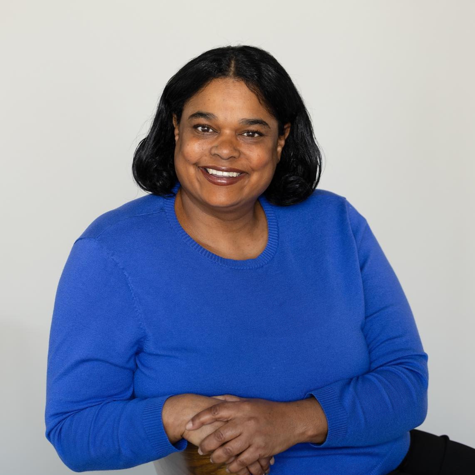 A woman with black hair, wearing a blue sweater, smiling and sitting with her hands clasped in front of her against a plain light gray background.