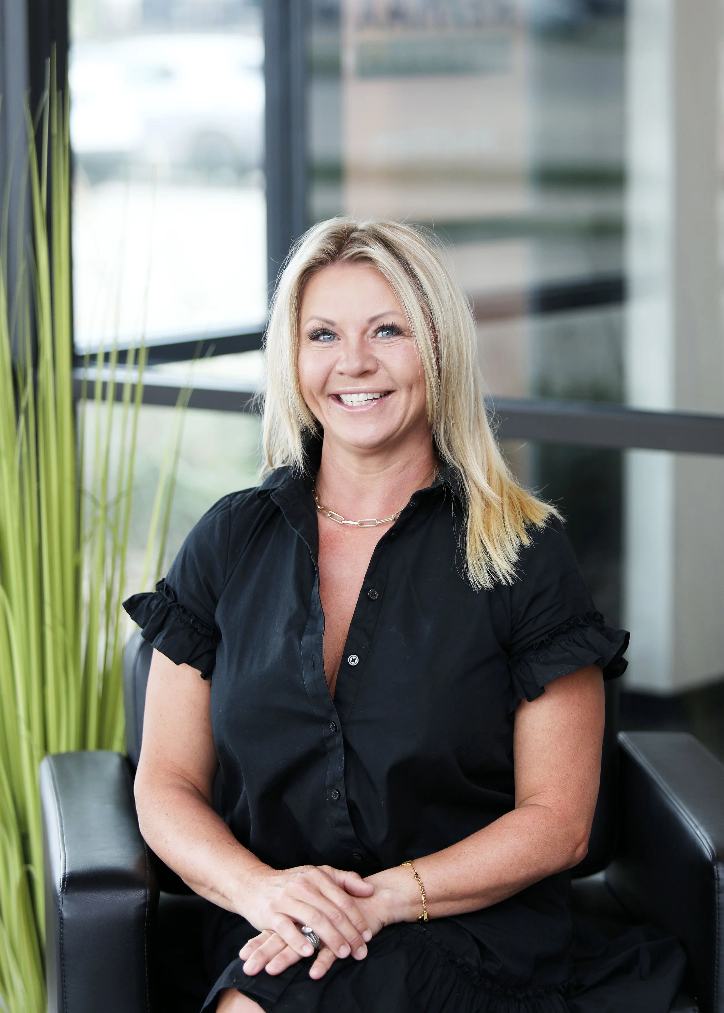 A smiling blonde woman wearing a black top and jewelry sitting in a modern office with large windows and greenery.