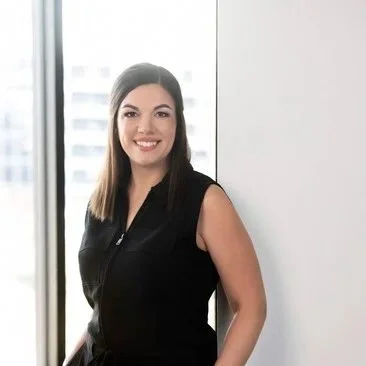 A woman with shoulder-length brown hair and a black sleeveless top smiling in front of a window in an office setting.