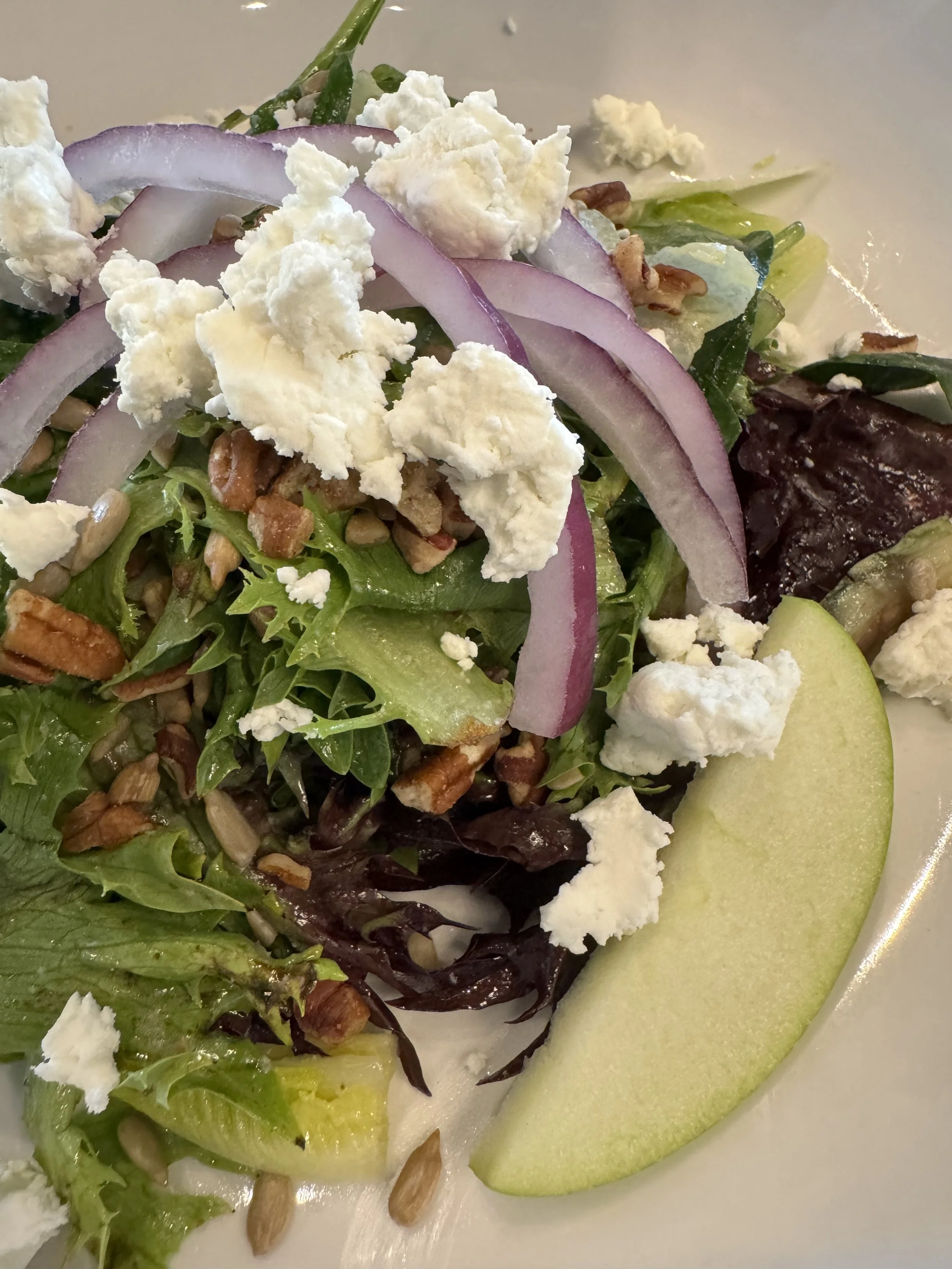 Close-up of a fresh mixed green salad topped with crumbled feta cheese, sliced red onions, chopped nuts, and sunflower seeds, with a slice of green apple on the side.
