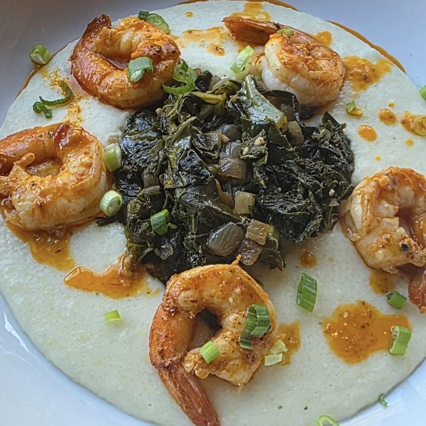 Creamy bowl with cooked shrimp, chopped green onions, and a pile of cooked dark leafy greens in the center, all garnished with a drizzle of orange sauce.