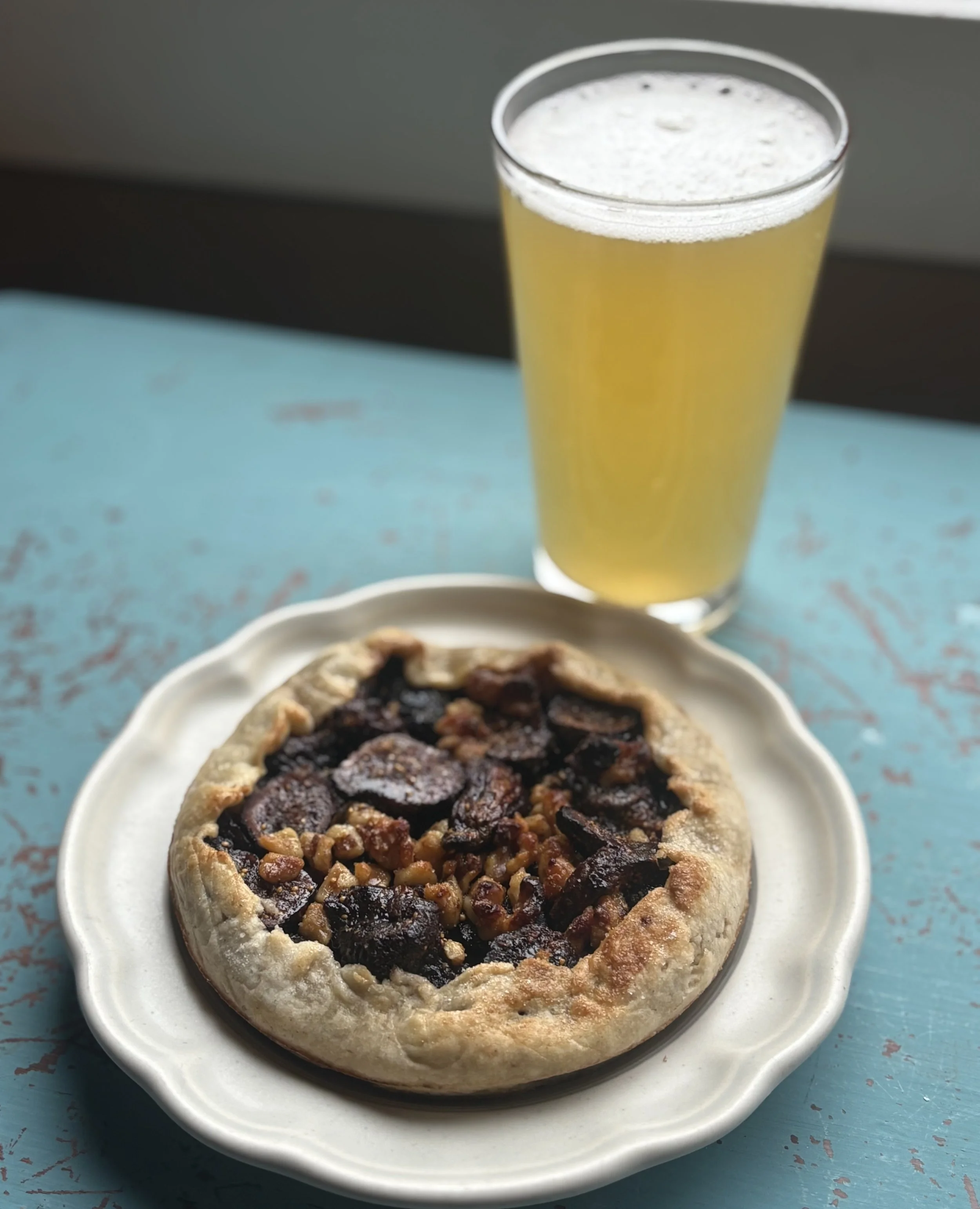 A dessert pizza with a nut topping served on a white plate and a tall glass of pale yellow beverage on a blue distressed table.