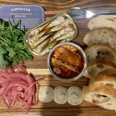 A wooden platter with slices of bread, pickled onions, fresh arugula, canned sardines, a can of anchovies, and a small bowl of tomato sauce.