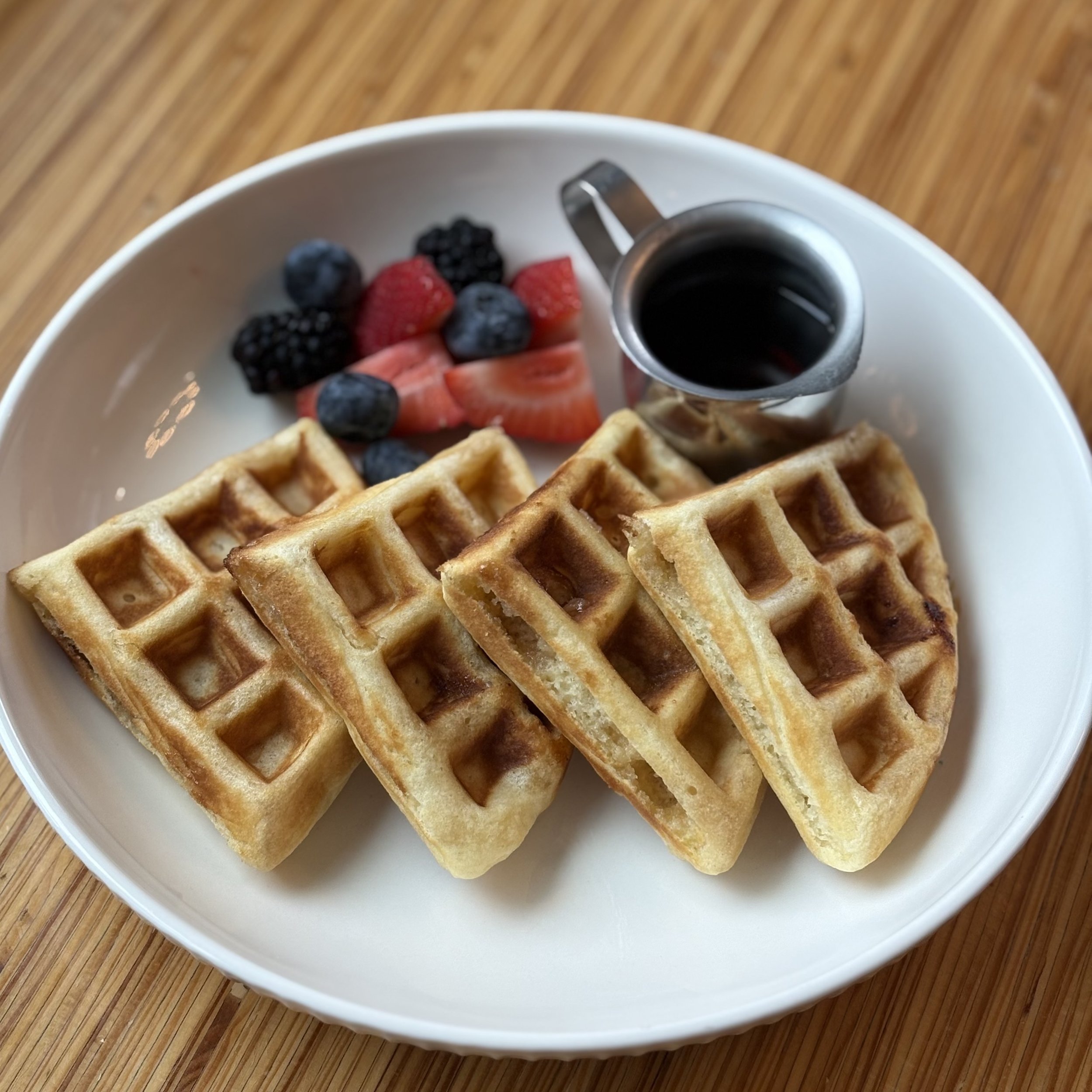 A white plate holding four waffles, a small pitcher of syrup, and a side of mixed berries including strawberries, blueberries, and blackberries, on a wooden table.