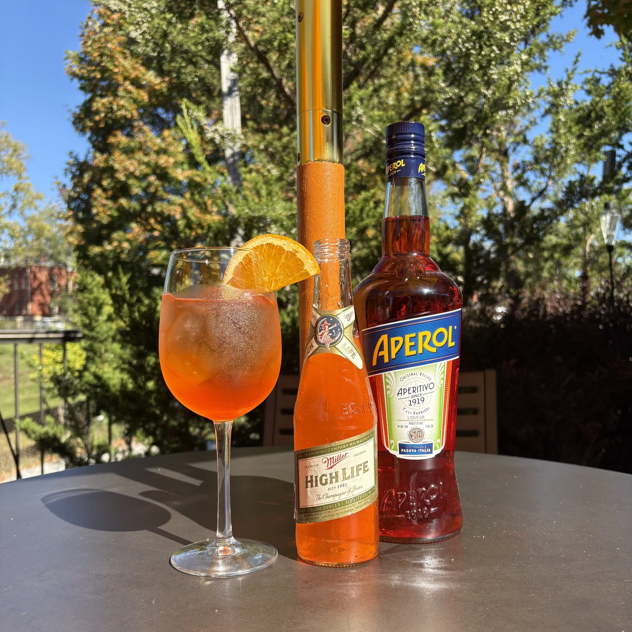 A glass of Aperol spritz with orange slice garnishing, a bottle of Aperol aperitif, and a bottle of Miller High Life beer on a gray outdoor table with trees and a blue sky in the background.