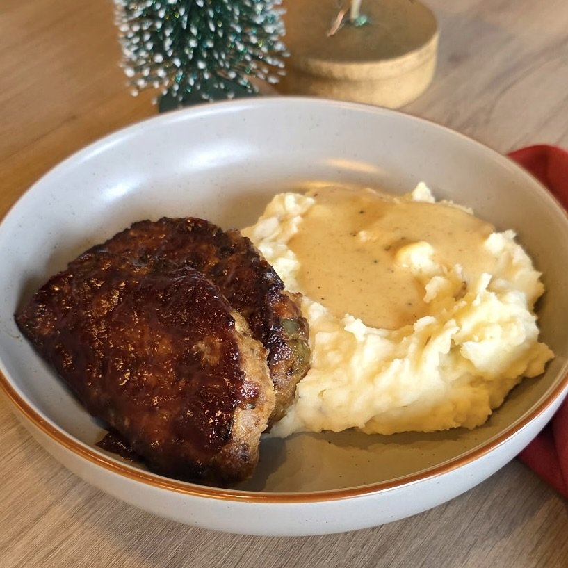 A bowl of mashed potatoes with gravy and a meatloaf on the side, garnished with a small Christmas tree in the background