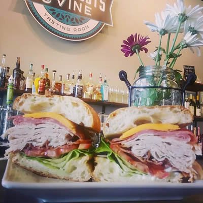 A sandwich cut in half revealing layers of lettuce, tomato, cheese, and deli meat on a plate, with a vase of white and purple flowers in the background.