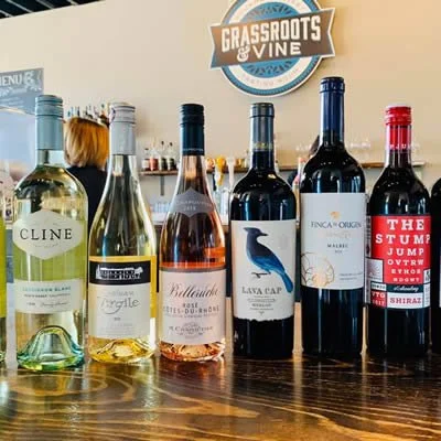 Six bottles of wine lined up on a wooden table with a 'Grassroots & Vine' sign in the background.