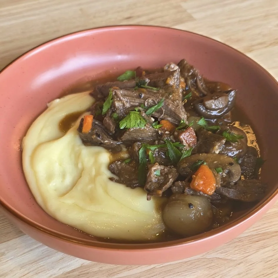 A bowl of creamy mashed potatoes topped with beef stew containing chunks of beef, carrots, mushrooms, and green herbs.