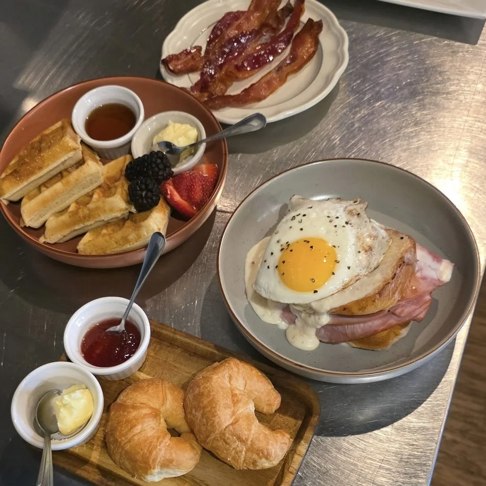 A breakfast spread includes a plate with a fried egg on top of ham and toast, a bowl of bacon with syrup, syrup and butter, a croissant on a wooden board with jam and butter, and a bowl of French toast with fresh berries.
