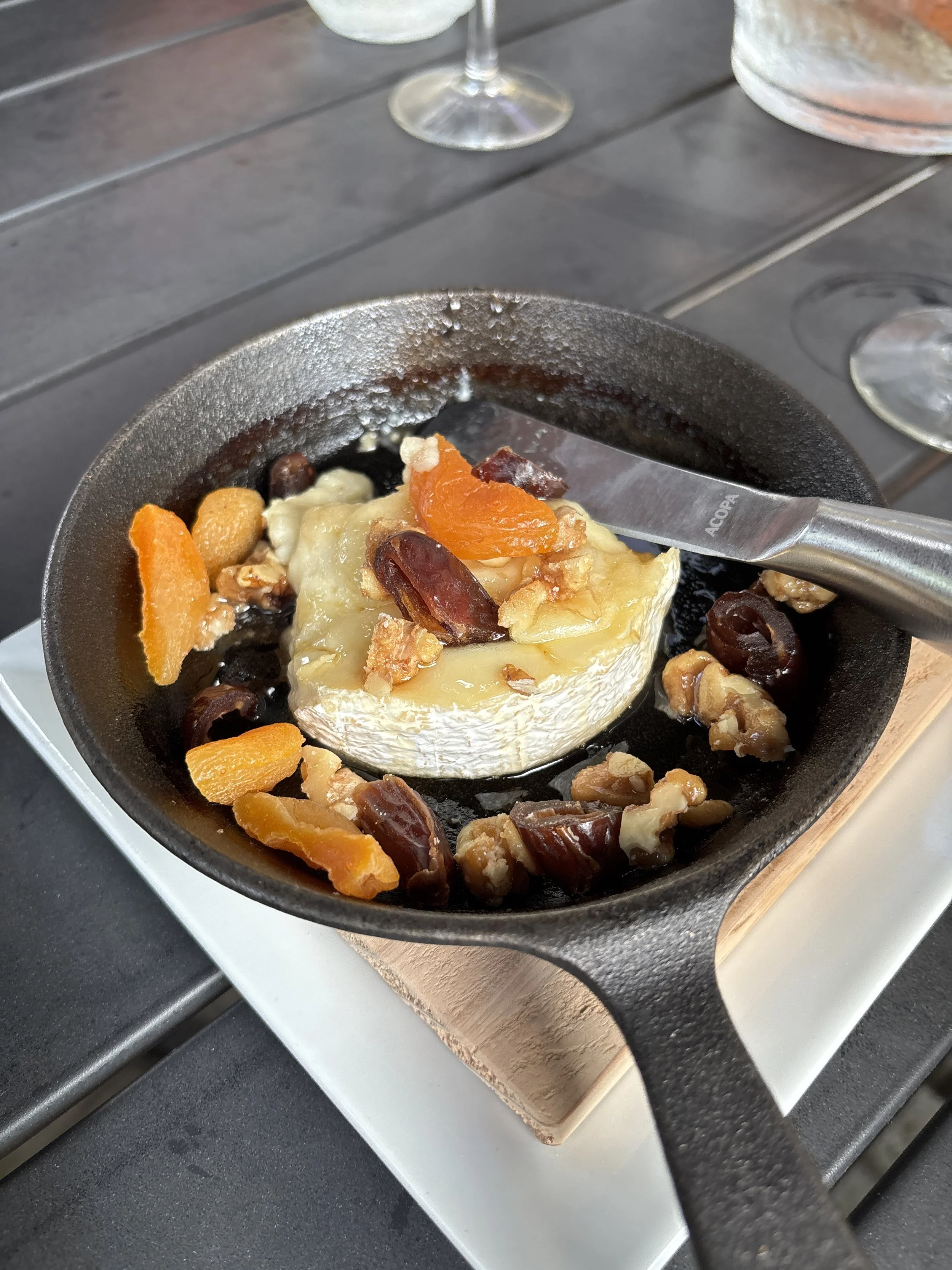 A small cast iron skillet with baked brie cheese topped with dried apricots, raisins, and walnuts, placed on a heating element. Two wine glasses are in the background on a dark wooden table.