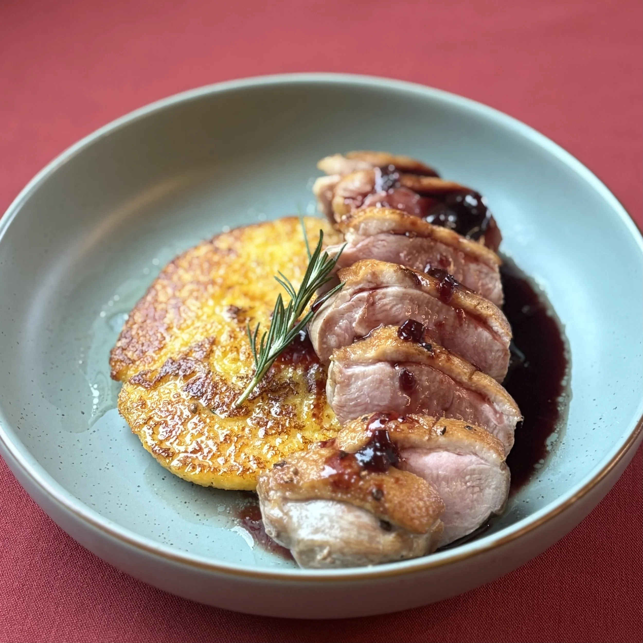 Seared duck breast sliced and topped with berry sauce, served with a golden hash brown garnished with a sprig of rosemary, on a light blue plate.