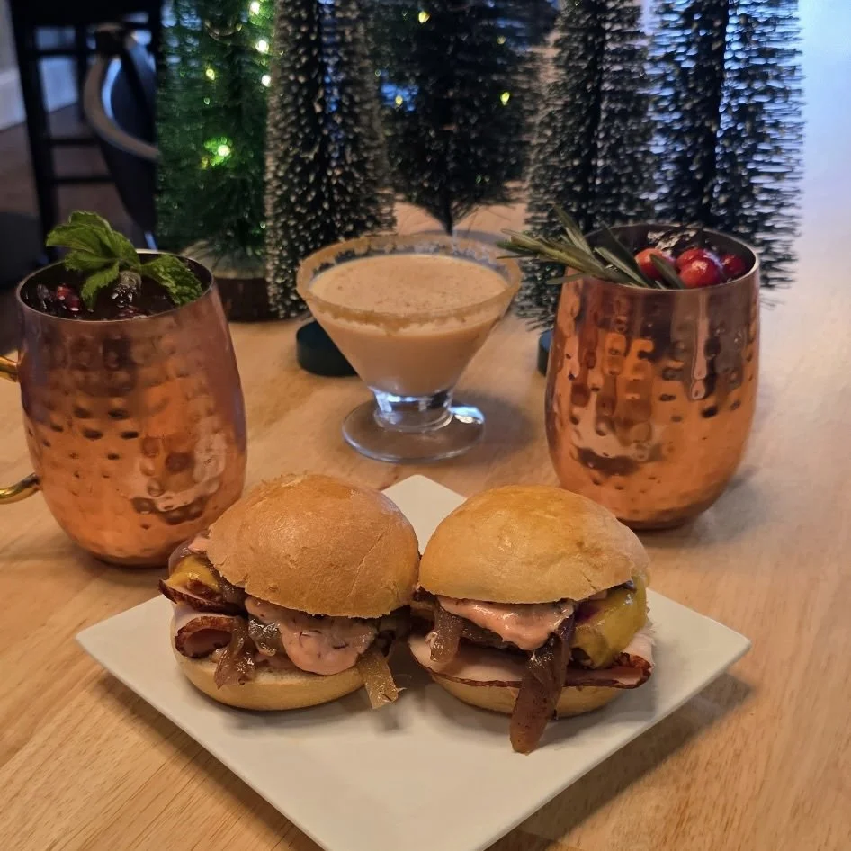 Two small sandwiches with beef, onions, and pickles on a white plate, two copper mugs with garnishes, and a creamy beverage in a glass goblet, with Christmas decorations in the background.