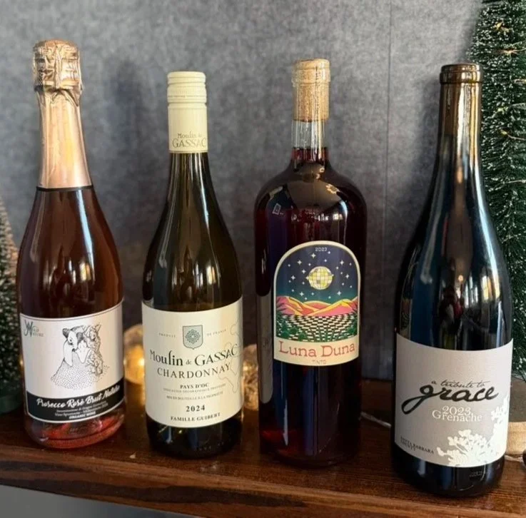 Four bottles of wine on a wooden shelf, with a gray background and a partial view of a small decorated Christmas tree on the right side.