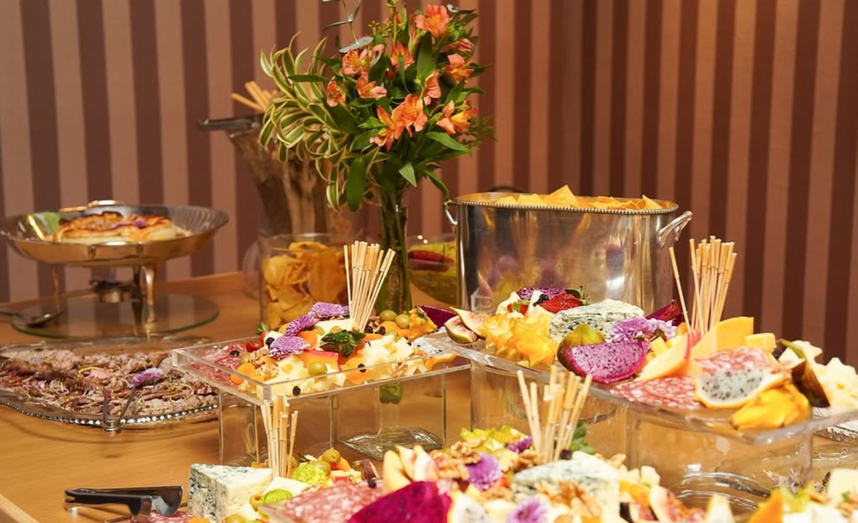 Mesa de festa com queijos, frutas, petiscos e uma planta decorativa no centro