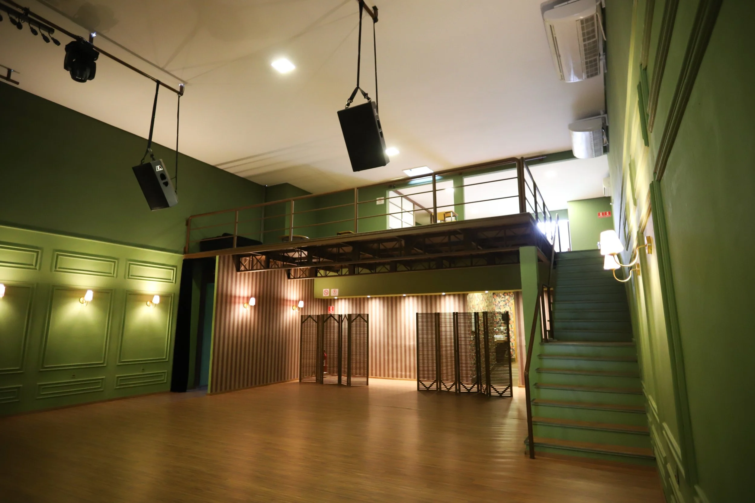 Salão de eventos com paredes verdes, piso de madeira, iluminação de parede, escadaria, área de palco com teto de metal, e sistema de som suspenso.