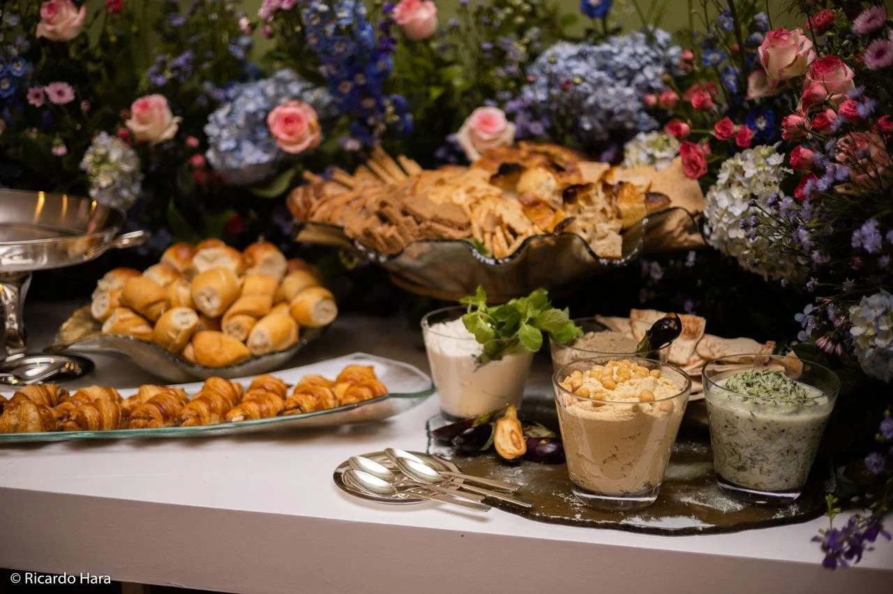 Mesa de brunch com várias opções de salgados, doces, pães, queijos, e três copos de creme ou patê, decorada com flores coloridas ao fundo.