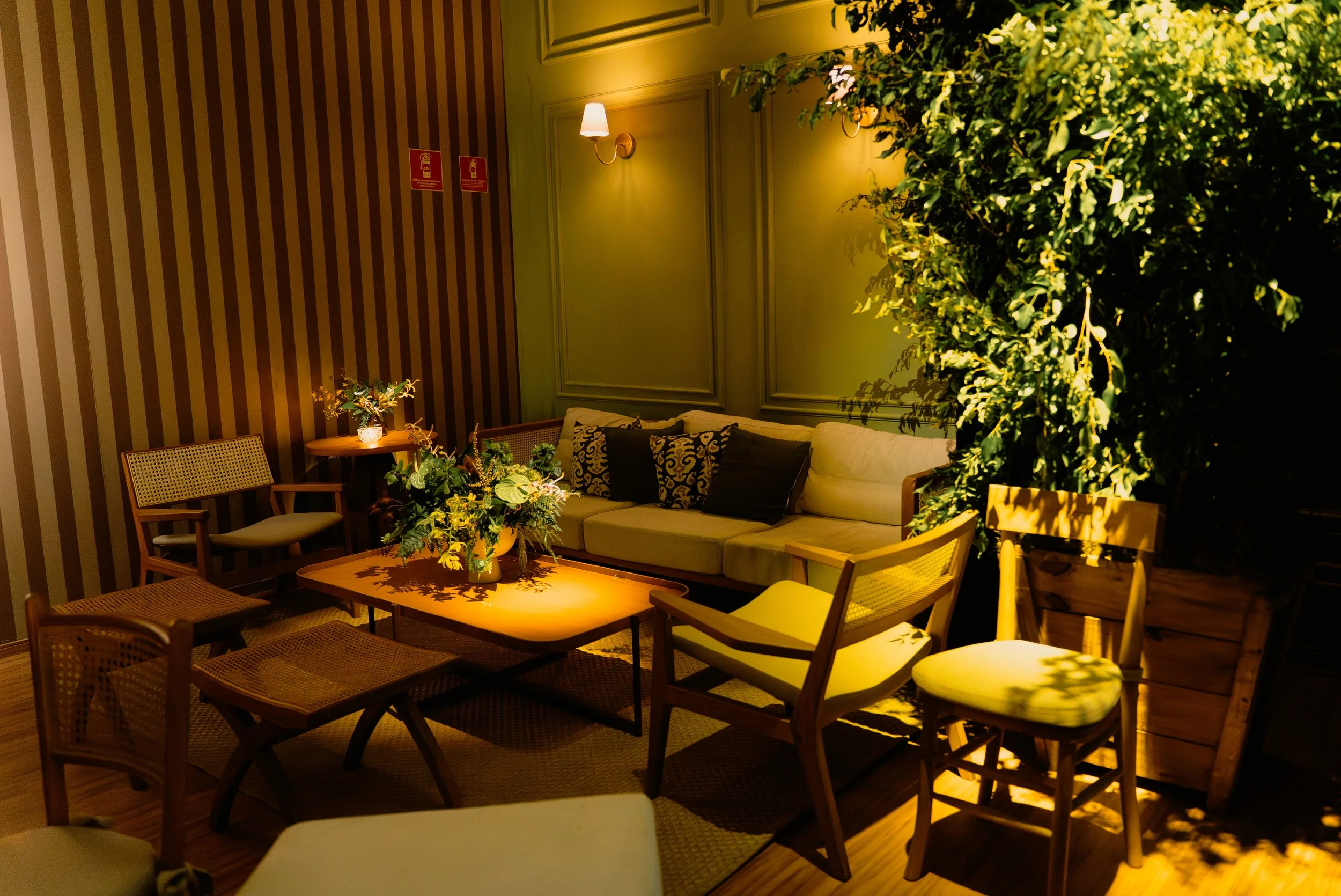 Sala de estar aconchegante com sofá no mezanino do espaço, cadeiras de madeira, mesa de centro, plantas e iluminação suave.