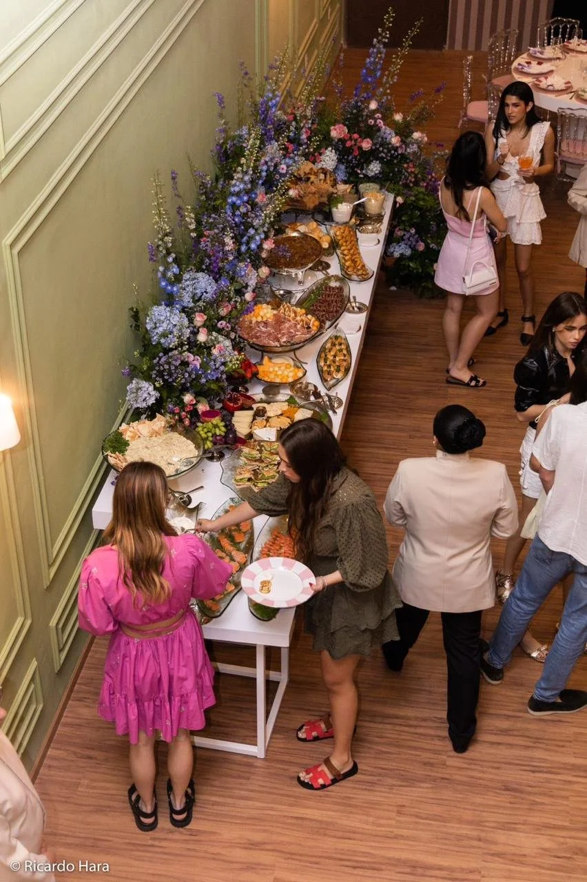 Pessoas servindo-se de comida em uma festa com buffet de alimentos variados, uma mesa decorada com flores e pratos decorativos, ambiente elegante com piso de madeira e paredes verdes.