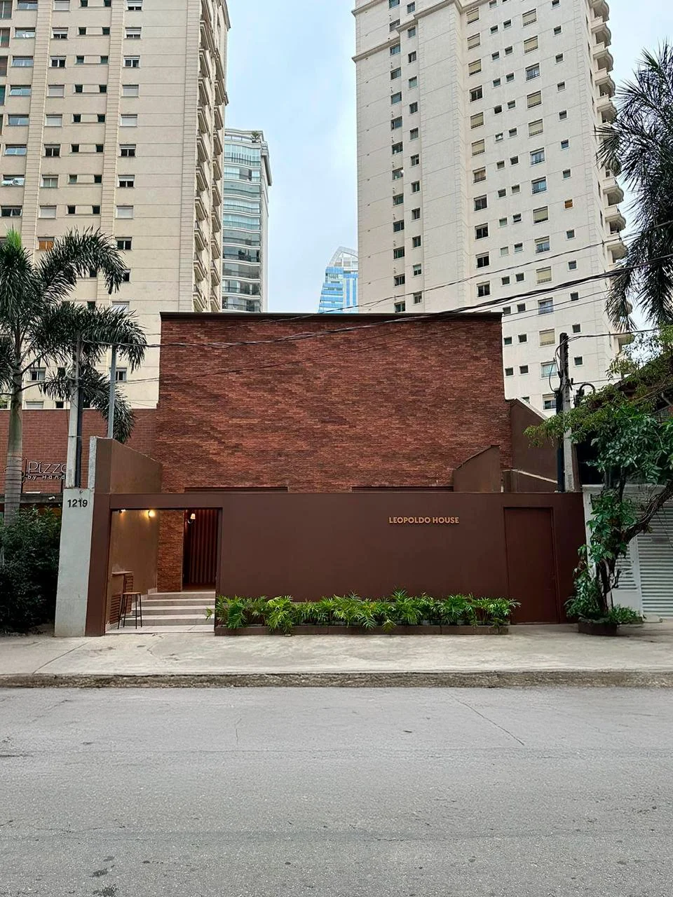 Edifício de fachada de parede de tijolos vermelhos com entrada marcada como 'Leopoldo House', com plantas na frente e prédios altos ao fundo.