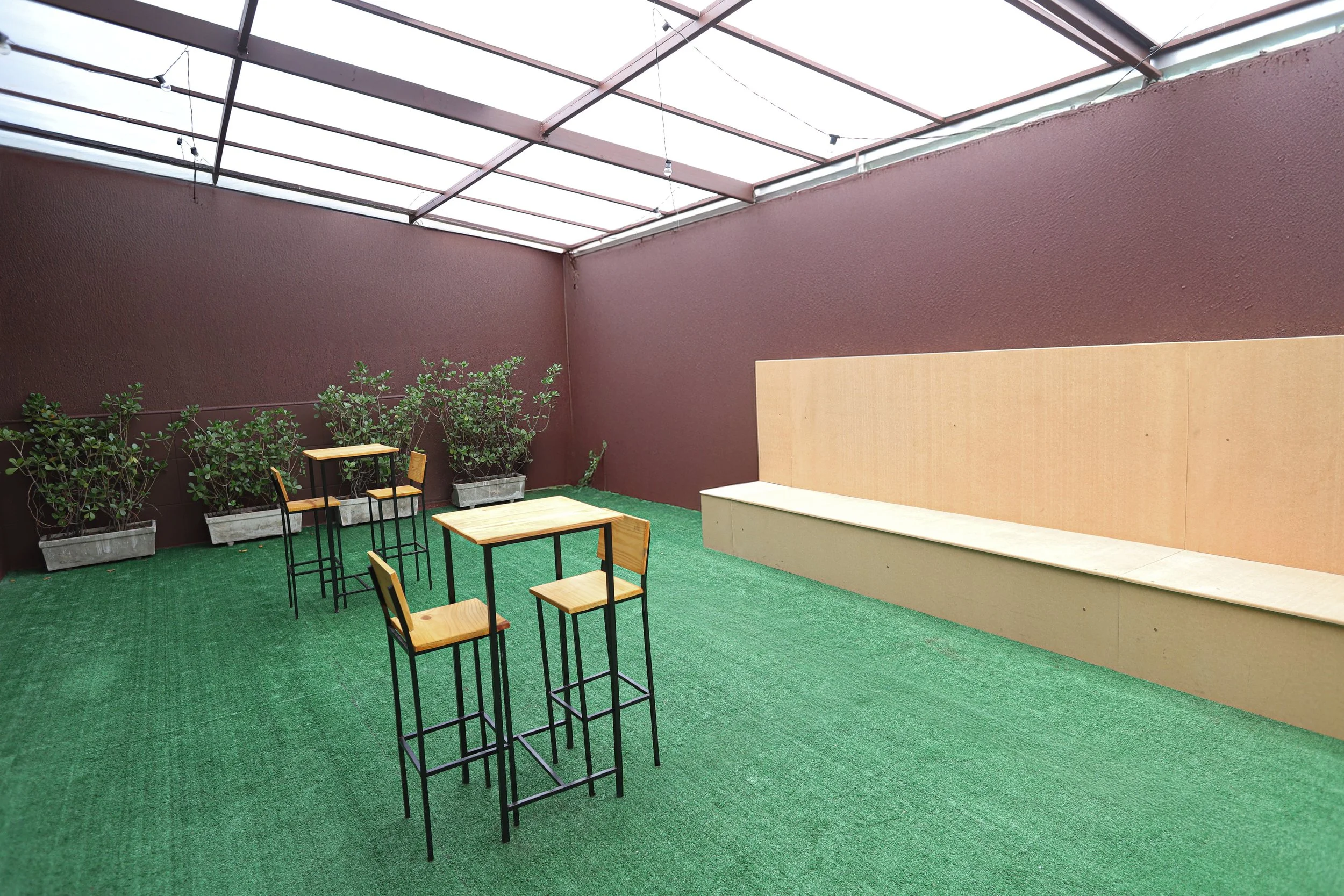 Rooftop Garden com varanda coberta com piso de grama artificial, com uma parede de cor vinho e plantas em vasos ao fundo. Há três mesas altas com cadeiras ao redor e uma parede de madeira ao lado.