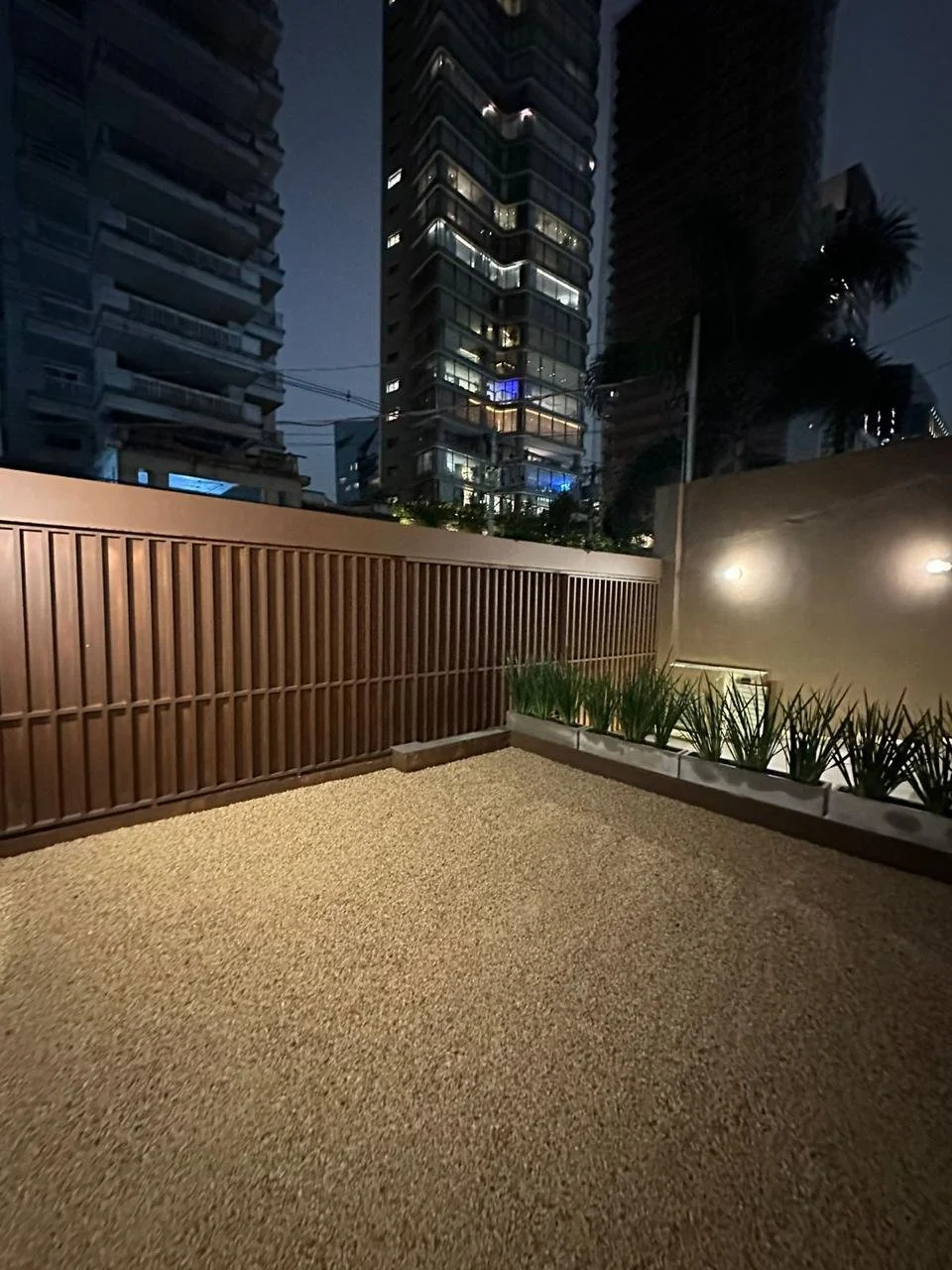 Varanda com piso de pedras, cerca de madeira, jardim com plantas e prédios altos ao fundo durante a noite.