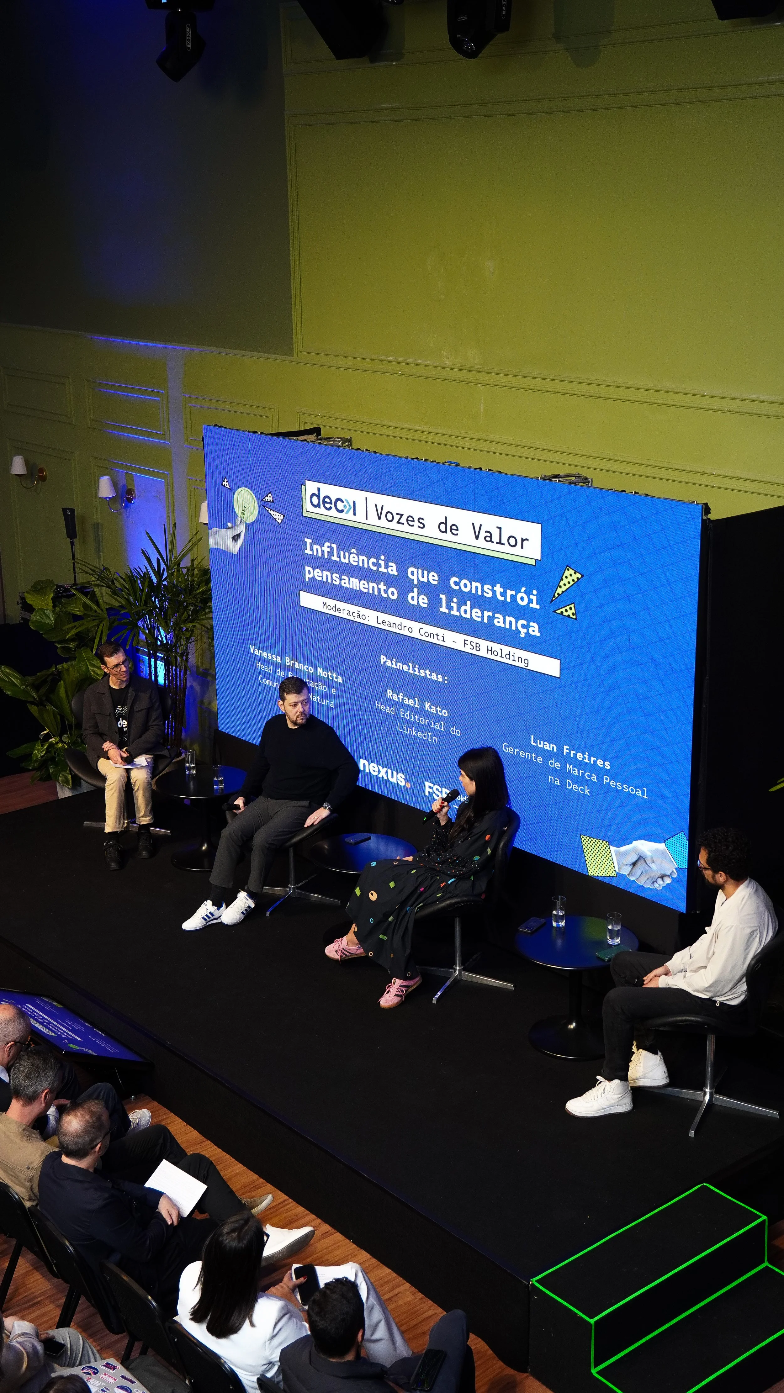 Painel de palestra com quatro pessoas sentadas no palco e público assistindo na plateia, com tela exibindo informações do evento, que é sobre liderança e influência, com nomes dos painelistas e moderador.