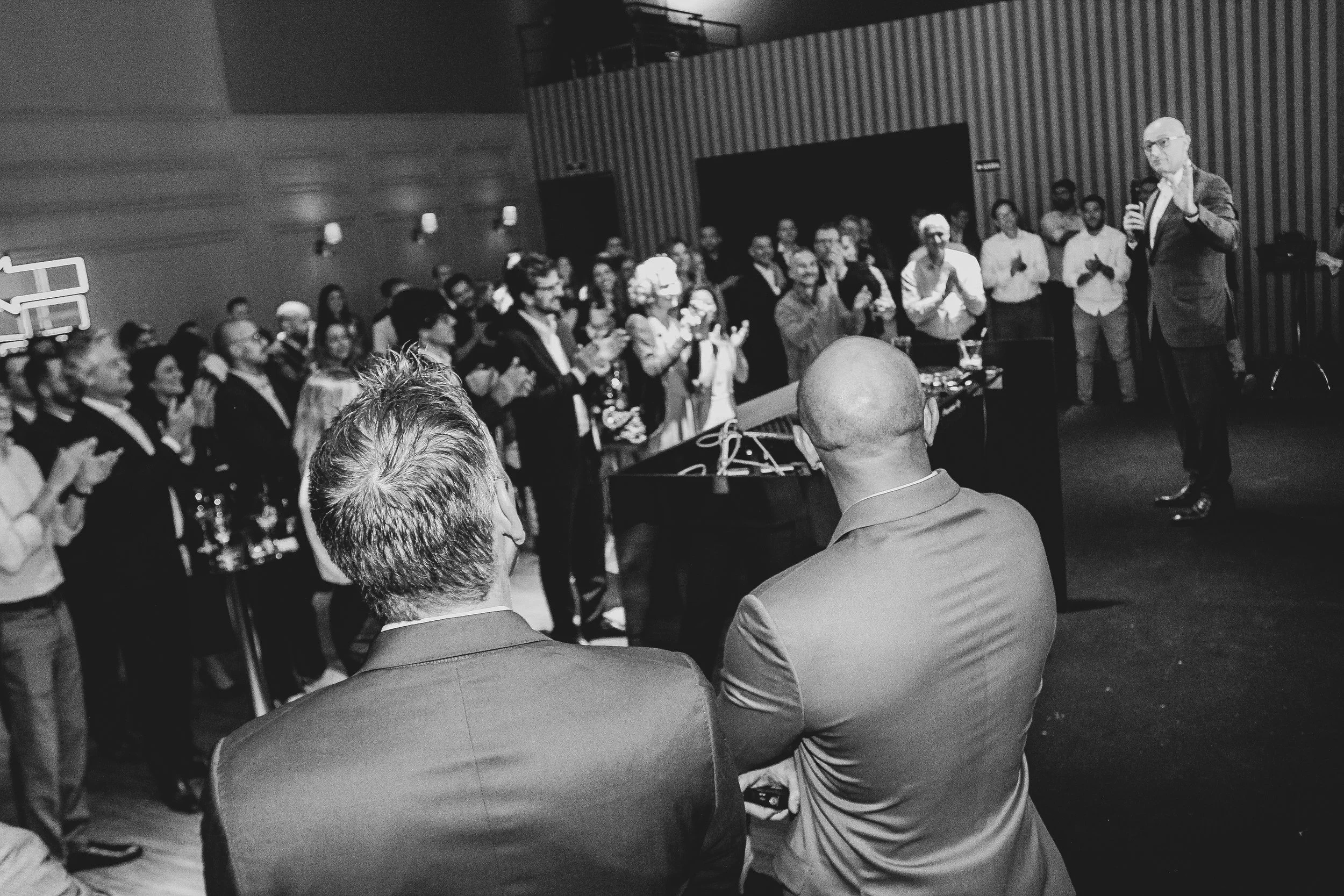 Homem de terno falando ao microfone em evento, com público assistindo e aplaudindo.