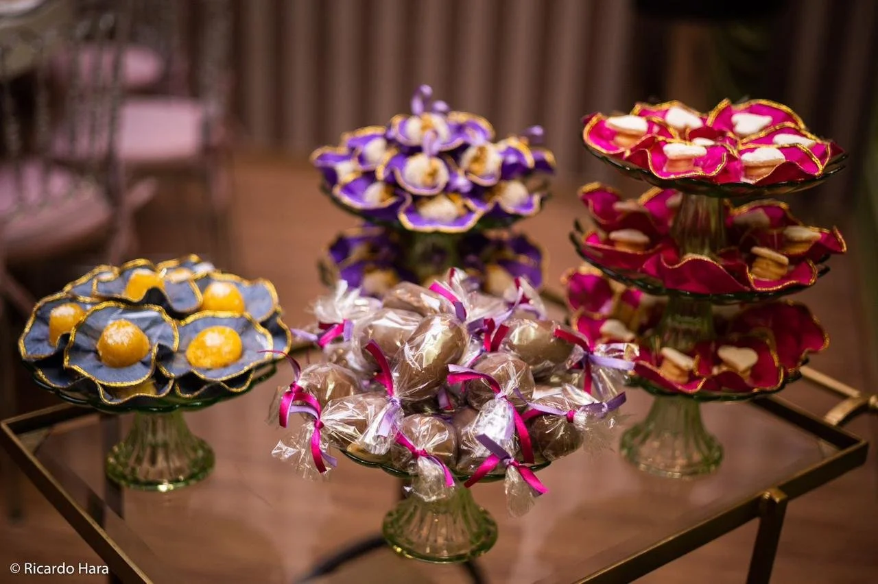 Doces de festa em diferentes cores, decorados com detalhes dourados, apresentados em suportes de vidro altos e dispostos em uma mesa de madeira.