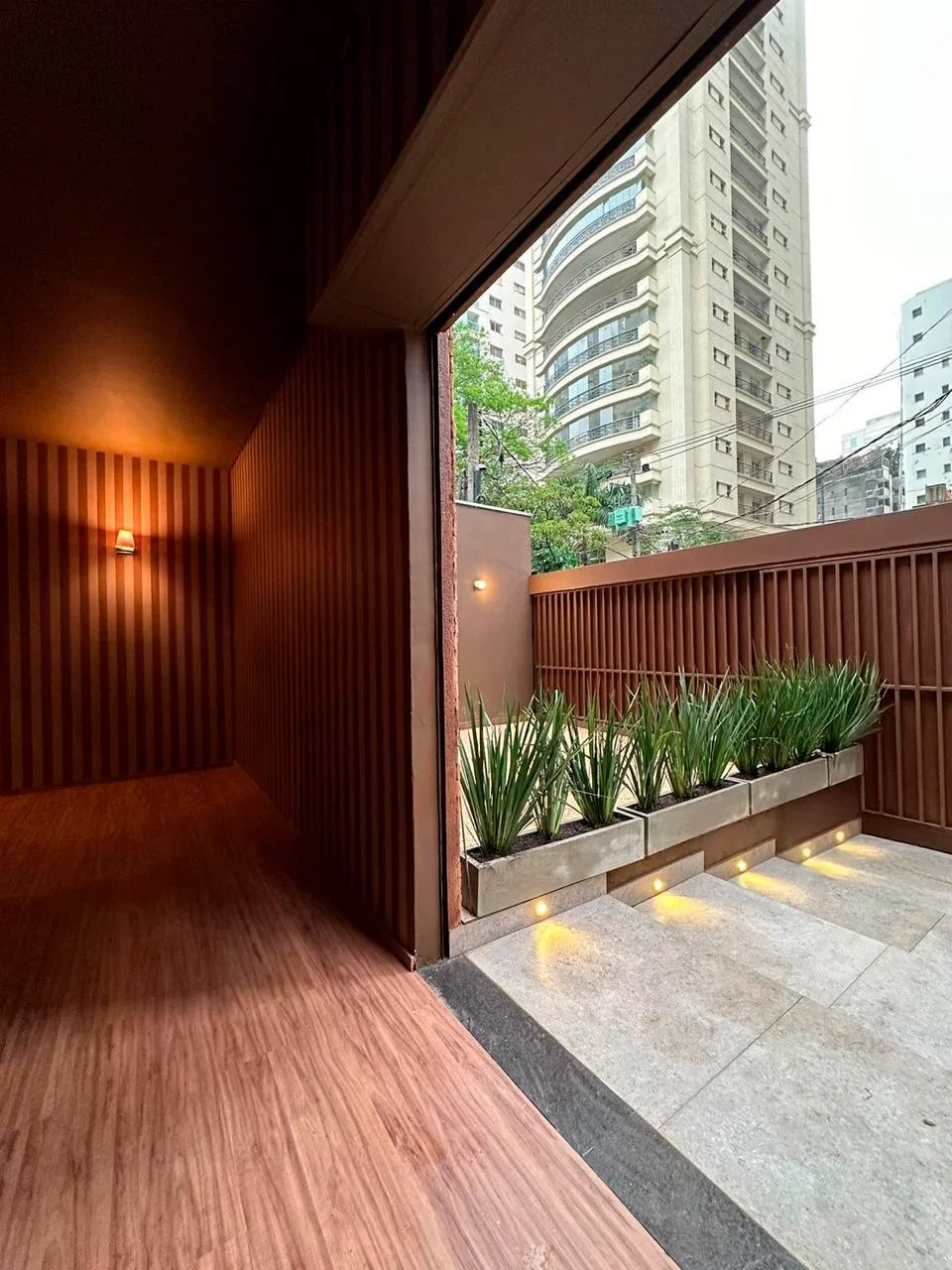 Apartamento com varanda, plantas em jardim vertical, piso de pedra, paredes de madeira com iluminação embutida, vista de prédios altos na cidade.