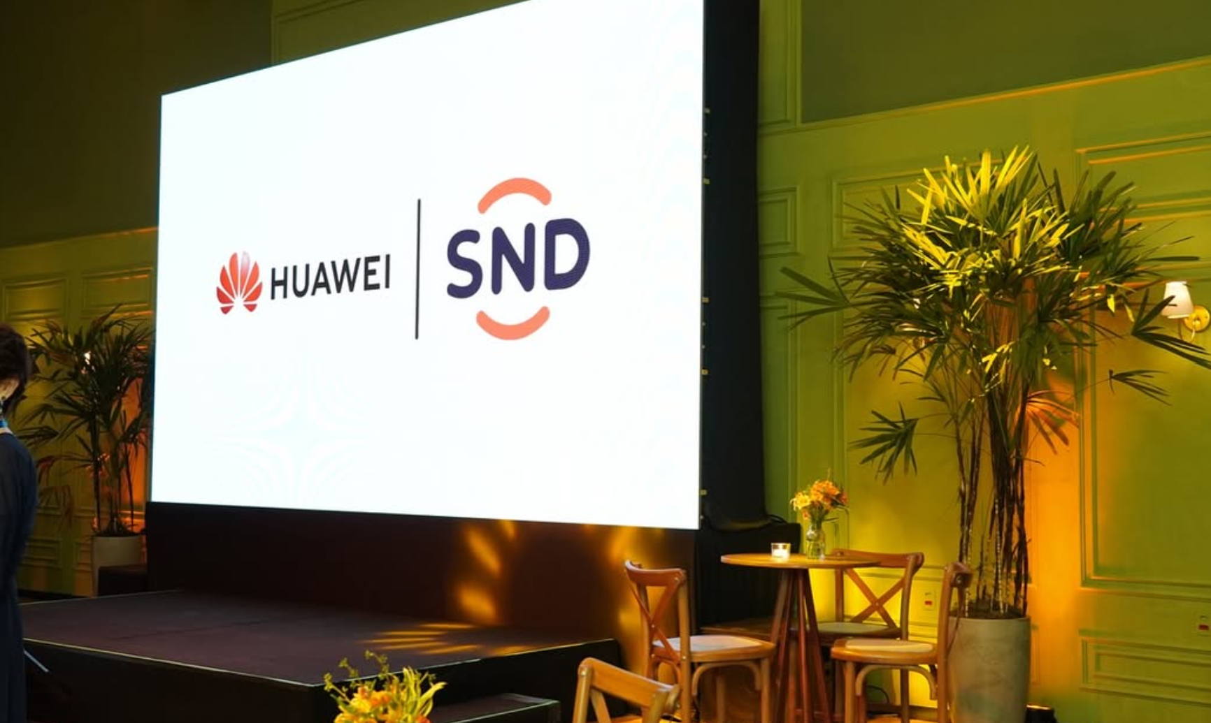 Tela e Painel de apresentação com os logotipos de marcas parceiras em evento corporativo B2B par alançamento de companha ou curso em um cenário decorado com plantas e iluminação amarelada.