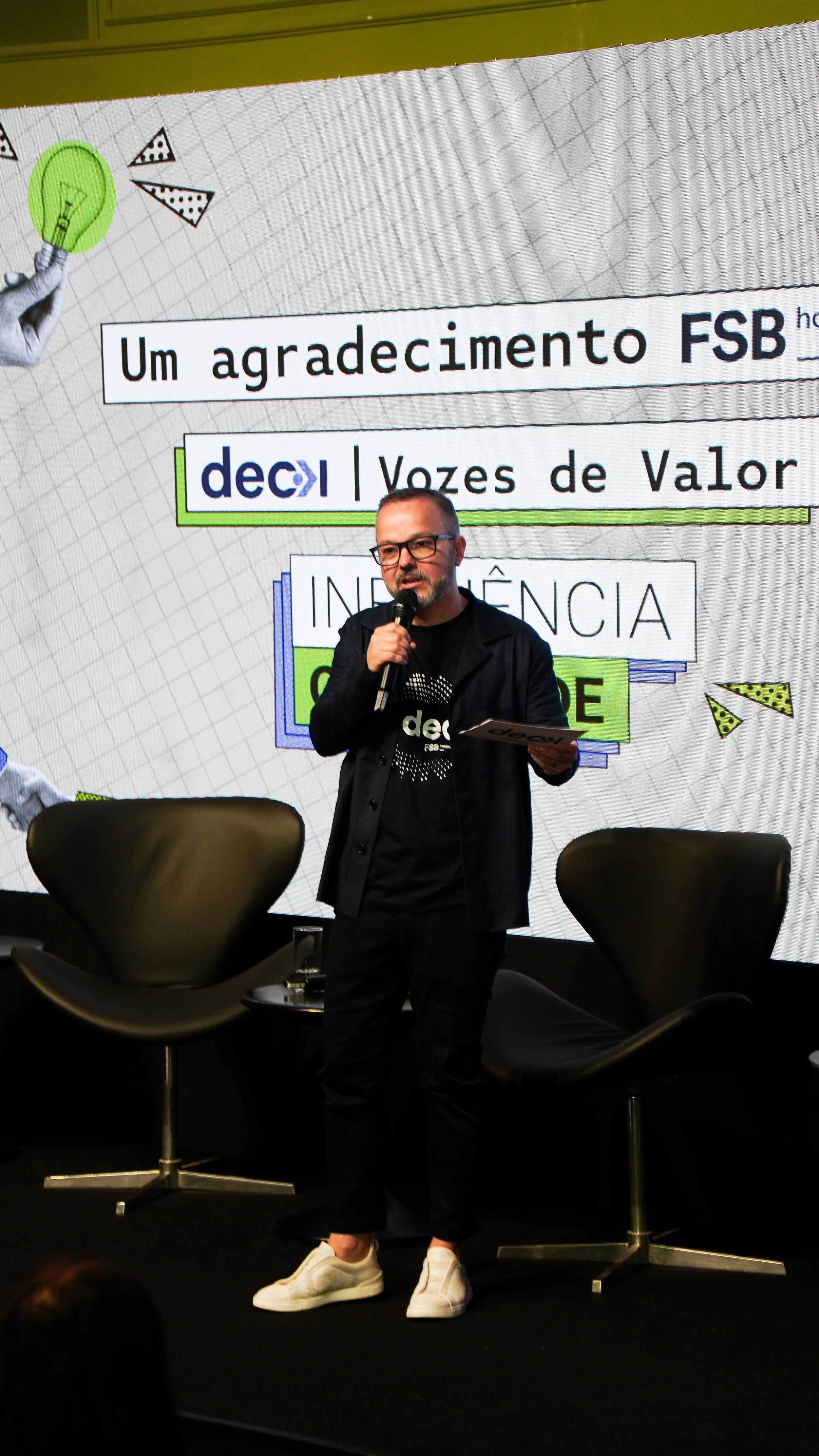 Homem em pé no palco segurando um microfone, vestindo jaqueta preta, t-shirt preta e tênis brancos. Atrás, uma tela com texto em português e elementos gráficos.