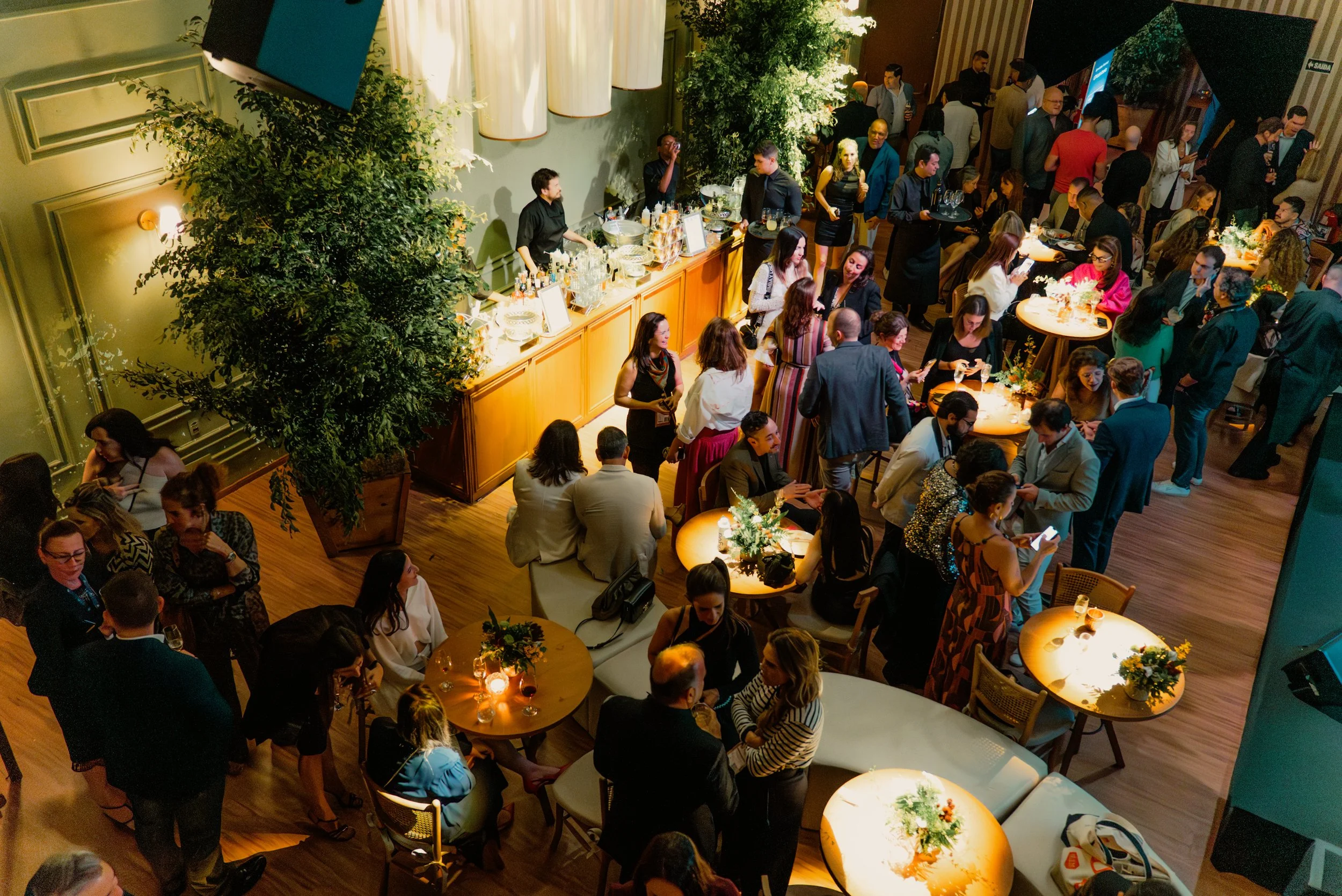 Pessoas socializando em um evento em um espaço elegante, com mesas decoradas com flores e um bar ao fundo, em um ambiente bem iluminado.