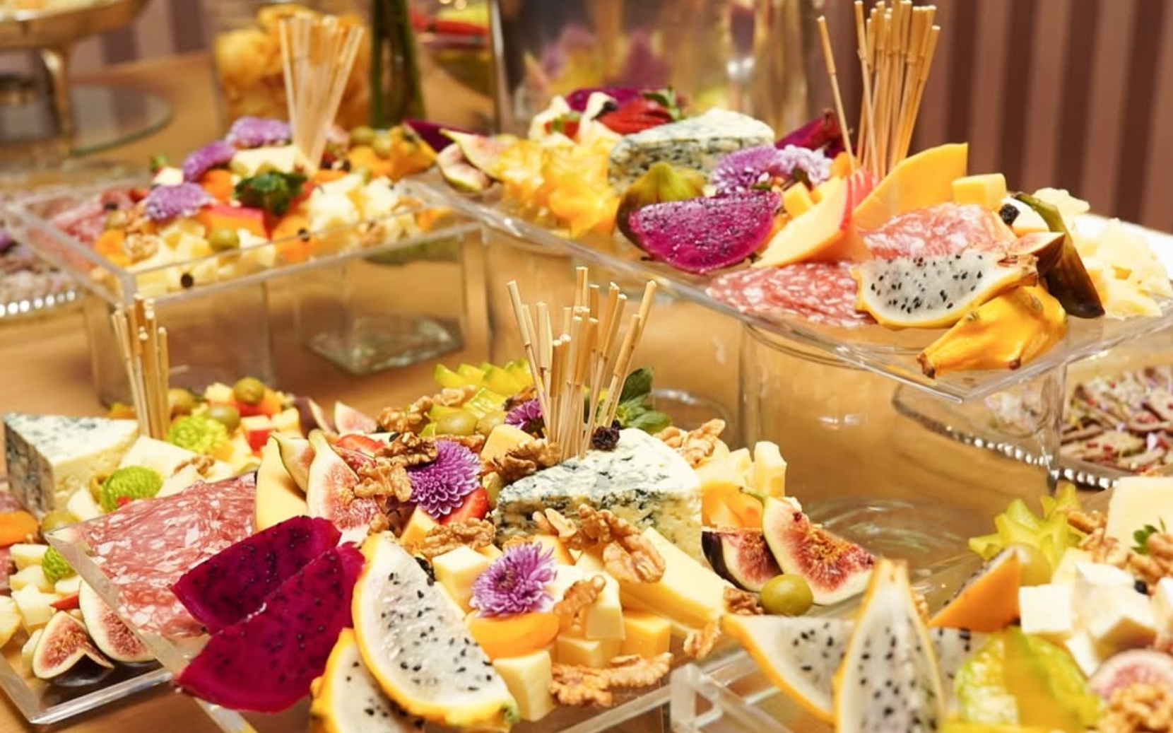 Mesas decoradas com diversas paletas de queijos, frutas frescas, nozes, geleias, e petiscos variados, apresentadas em pratos e suportes de vidro e acrílico.