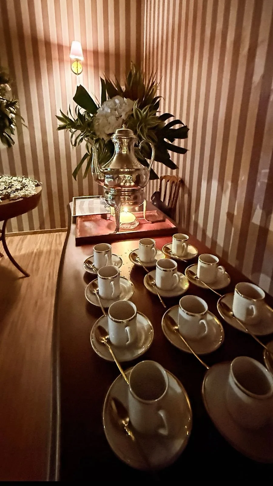 Mesa decorada com xícaras de café, pires e colheres de colher de prata, com um vaso com plantas e uma jarra de prata ao fundo e uma parede de listras rosa e bege ao fundo.