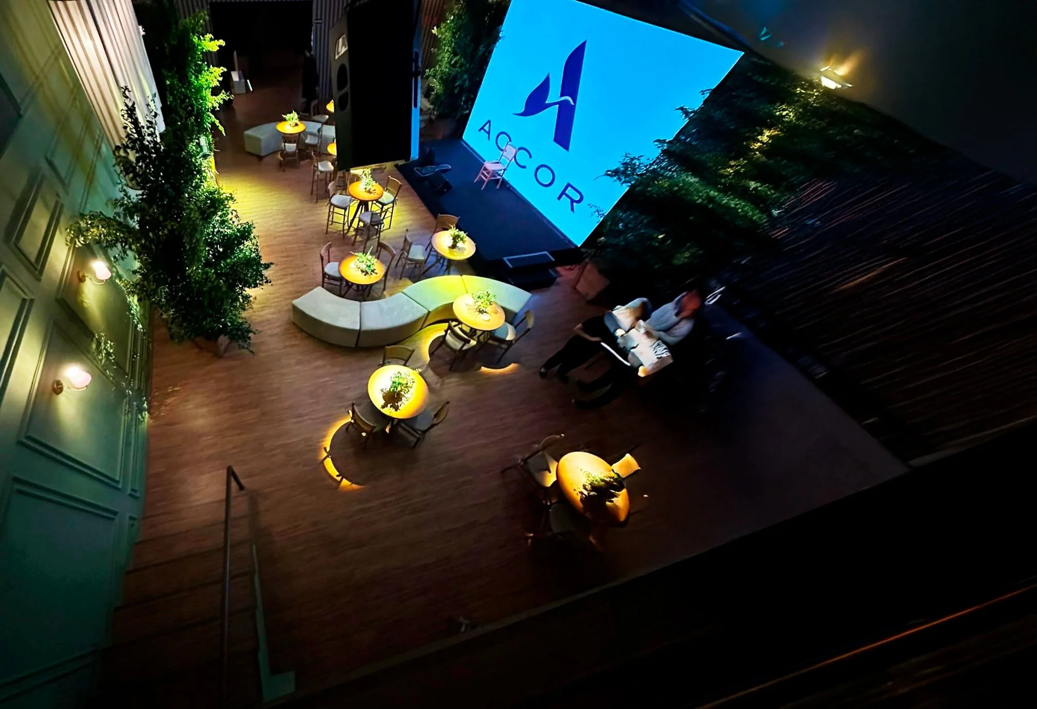 Sala de eventos com mesas redondas, uma tela grande exibindo o logotipo da Accor, cadeira e piano em destaque, iluminação suave, plantas decorativas e duas pessoas sentadas em um banco.