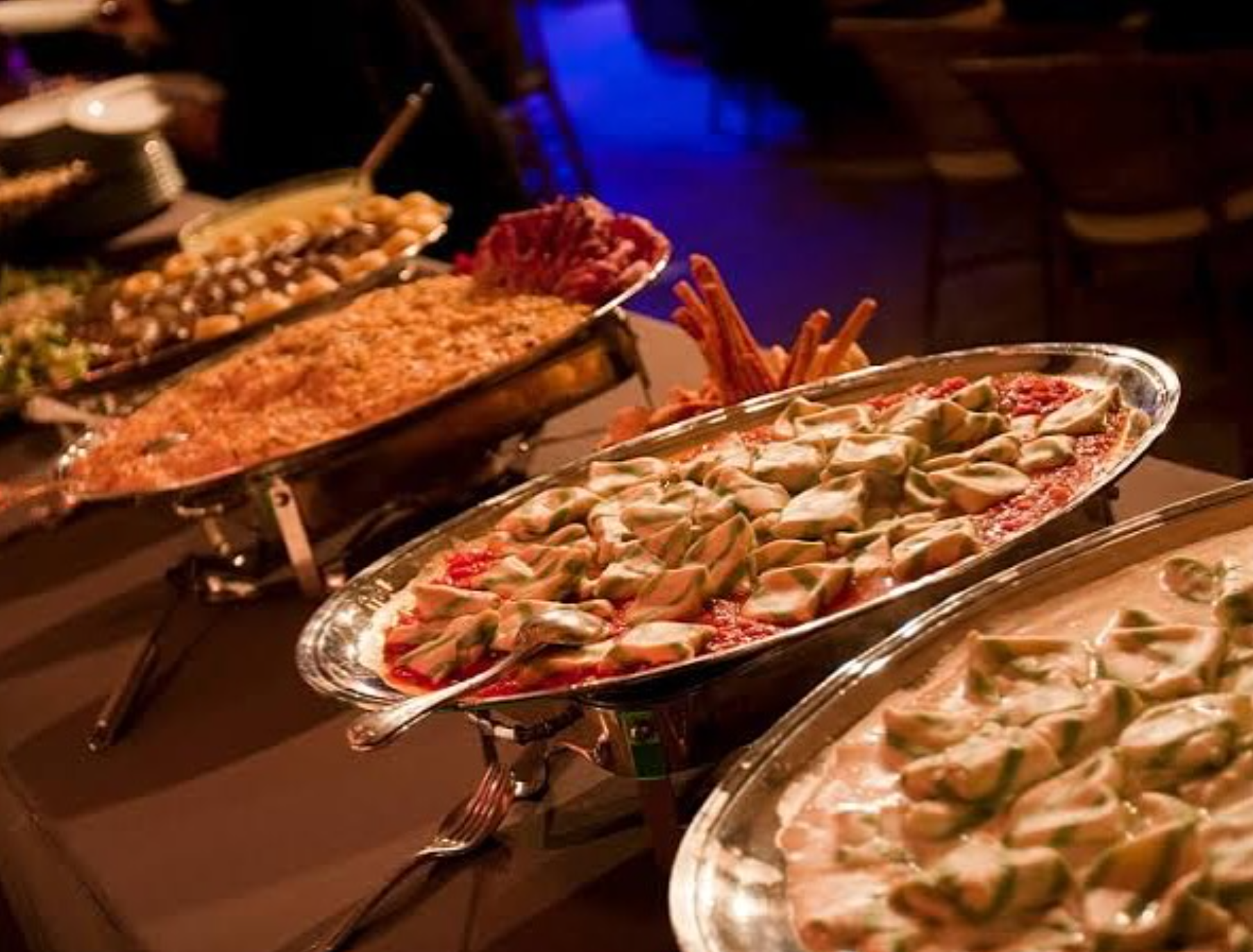 Buffet de comida com diferentes pratos, incluindo massas, tomates, acompanhamentos e entradas, em um ambiente com iluminação quente.