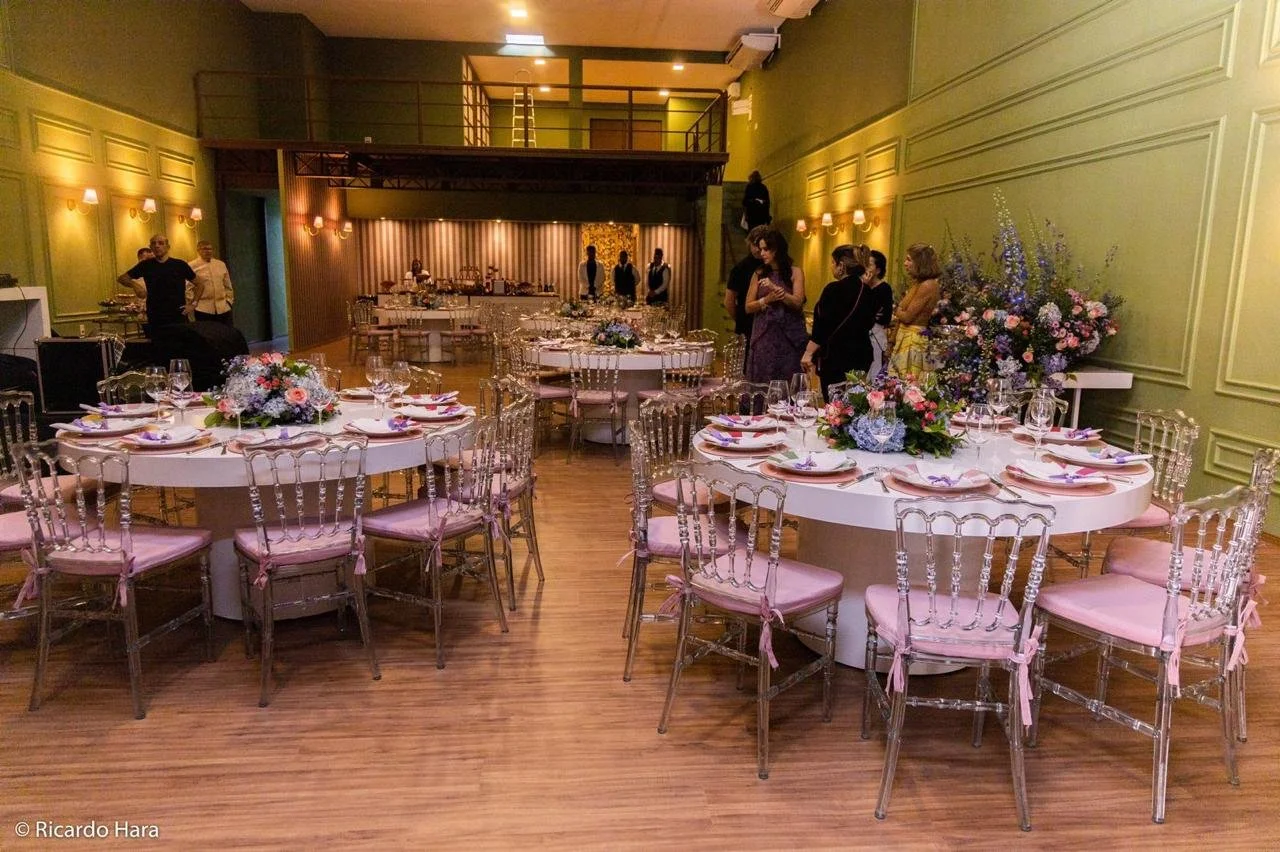 Salão de festa decorado com mesas redondas com flores e chácaras transparentes, cadeiras rosas com laços. Pessoas conversando ao fundo, paredes verdes com iluminação amarelada.