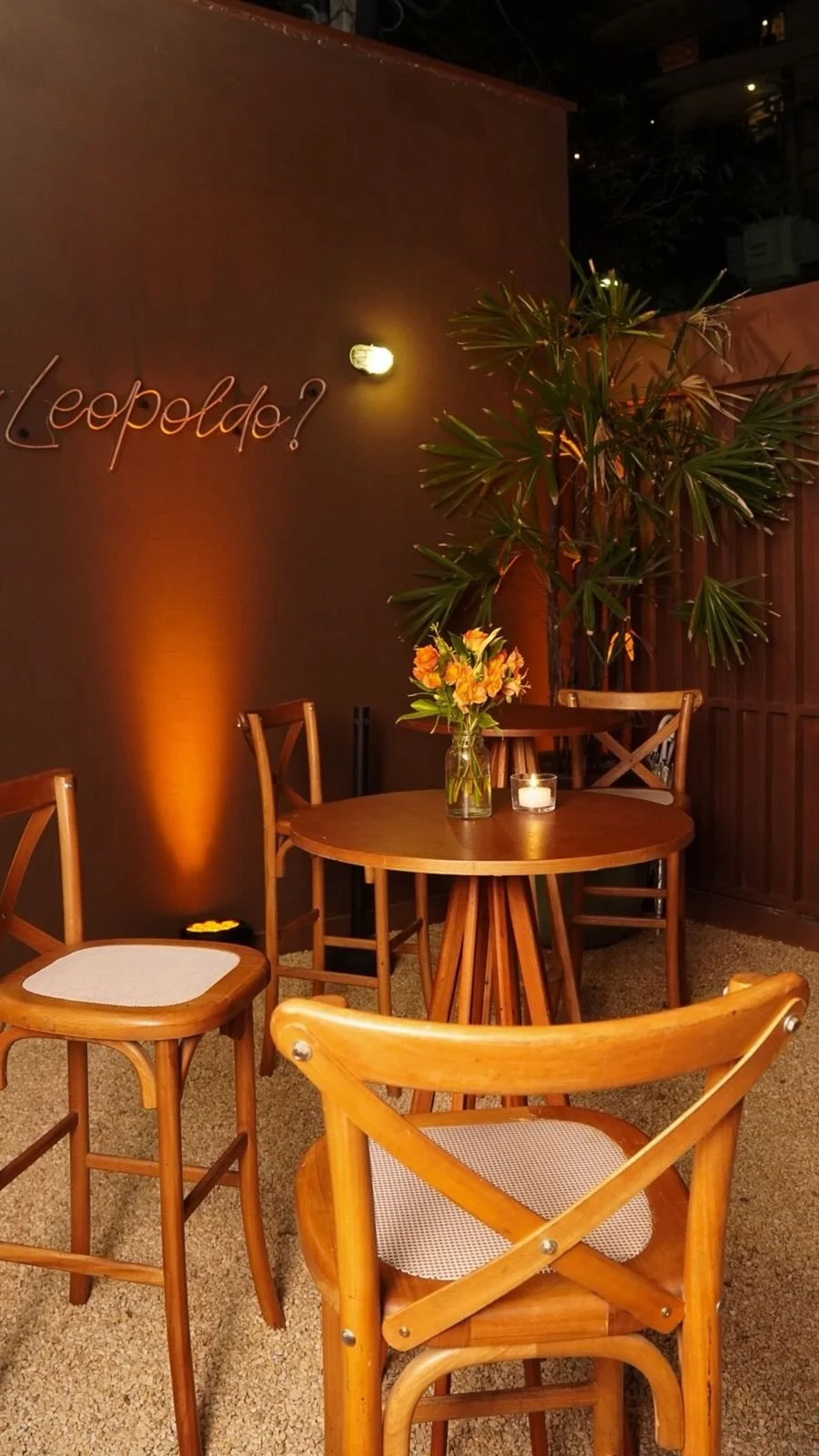 Área externa do espaço conhecida como praia ou prainha ao ar livre com: mesa de madeira, cadeiras de madeira, vaso com flores, vela acesa, parede com luz suave e letreiro em neon que diz " Who is Leopoldo?".