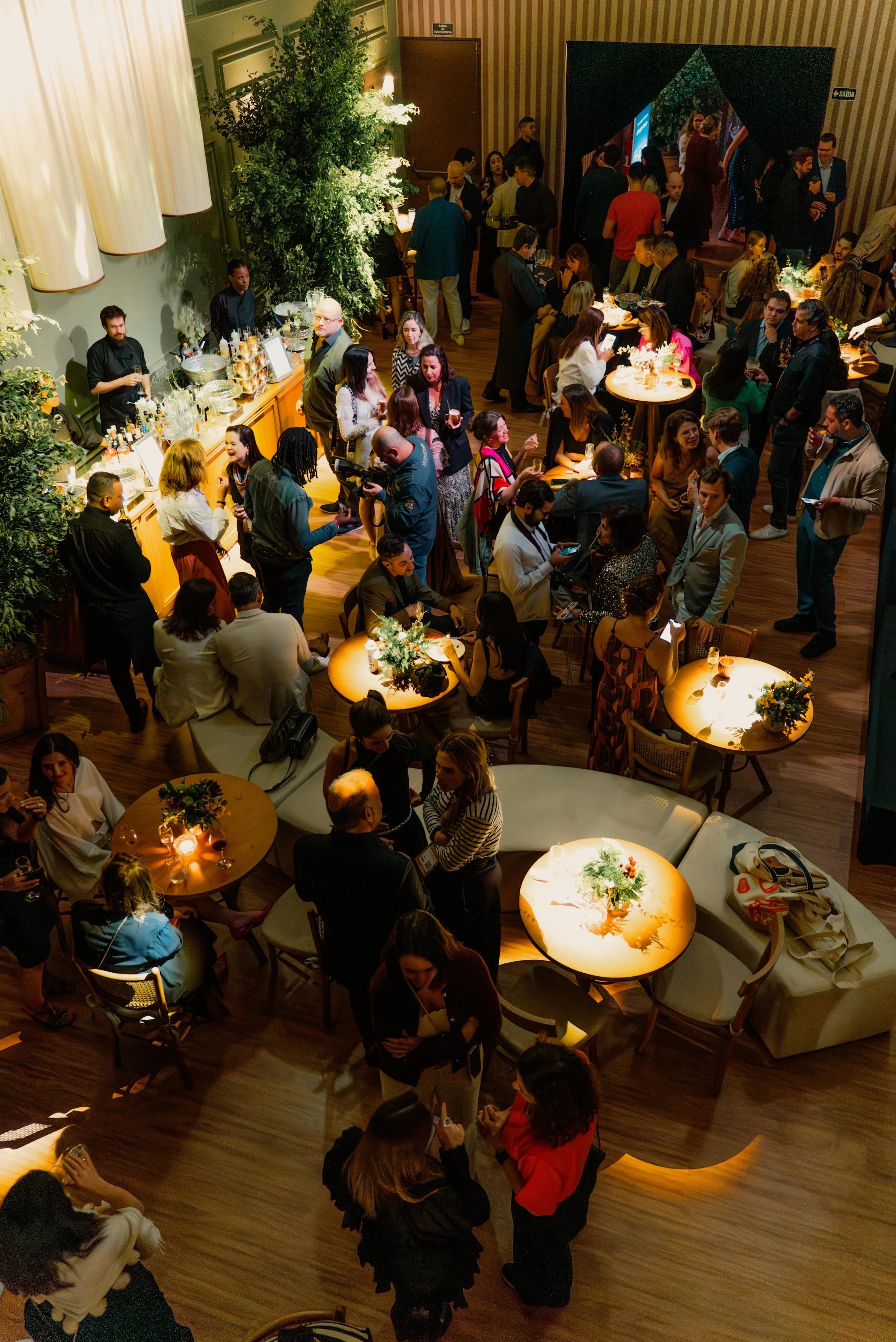 Cena de uma festa ou evento social com muitas pessoas interagindo, algumas sentadas e outras em pé, com mobiliário elegante, iluminação quente e decoração com flores e plantas. Pé direito de 7 metros o que torna o espaço mais arejado e amplo.